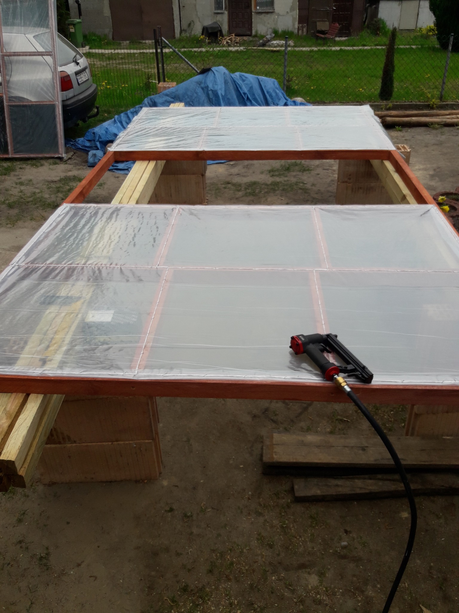 Konstrukcja drewniana z przezroczystym pokryciem, mocowana zszywaczem pneumatycznym, w tle widoczny samochód i trawnik.