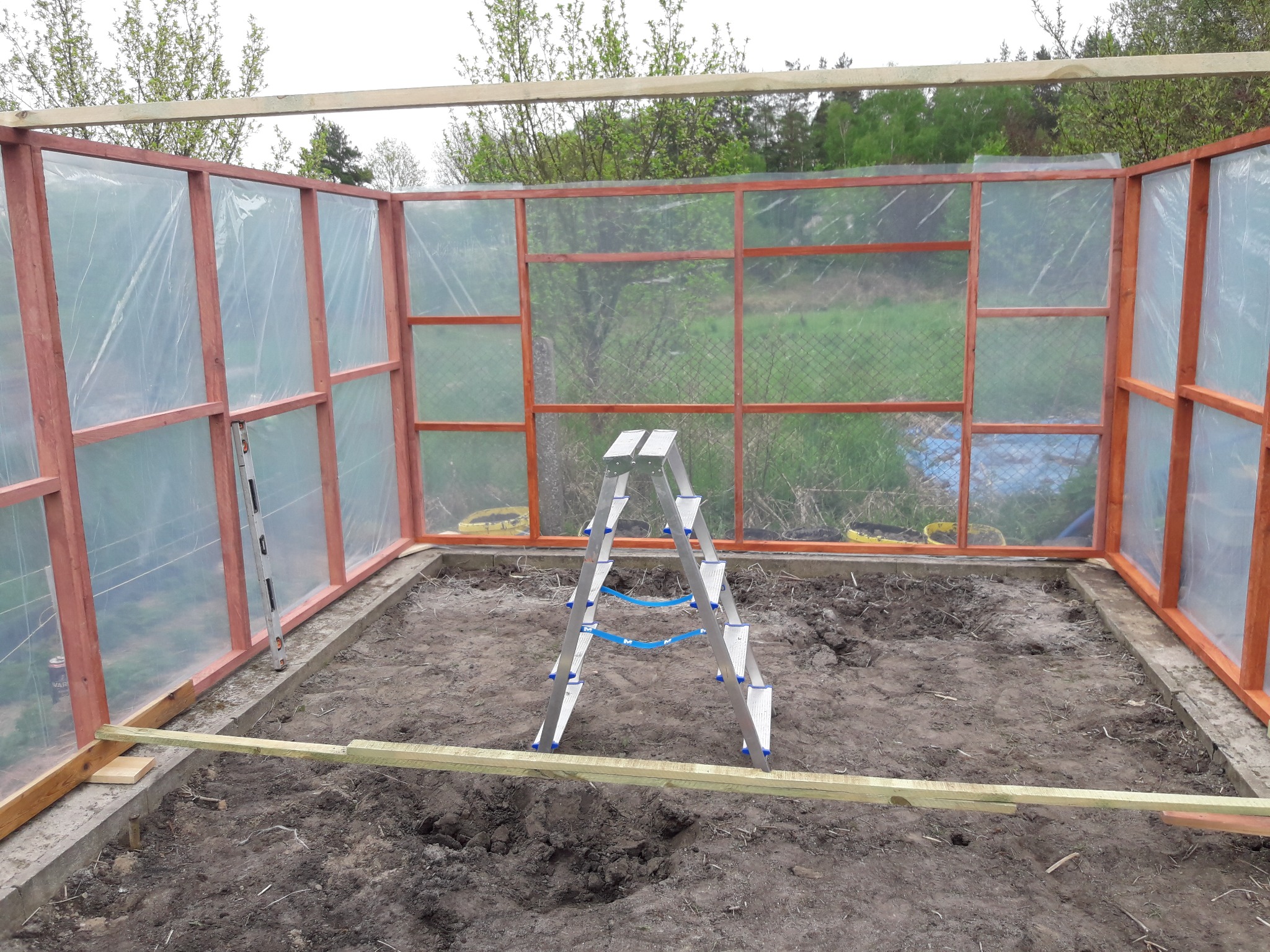 Konstrukcja szklarni w trakcie budowy: drewniany szkielet z folią, aluminiowa drabina na ziemi wewnątrz, poziomica przymocowana do ramy, widok na zewnątrz przez przezroczyste ściany.