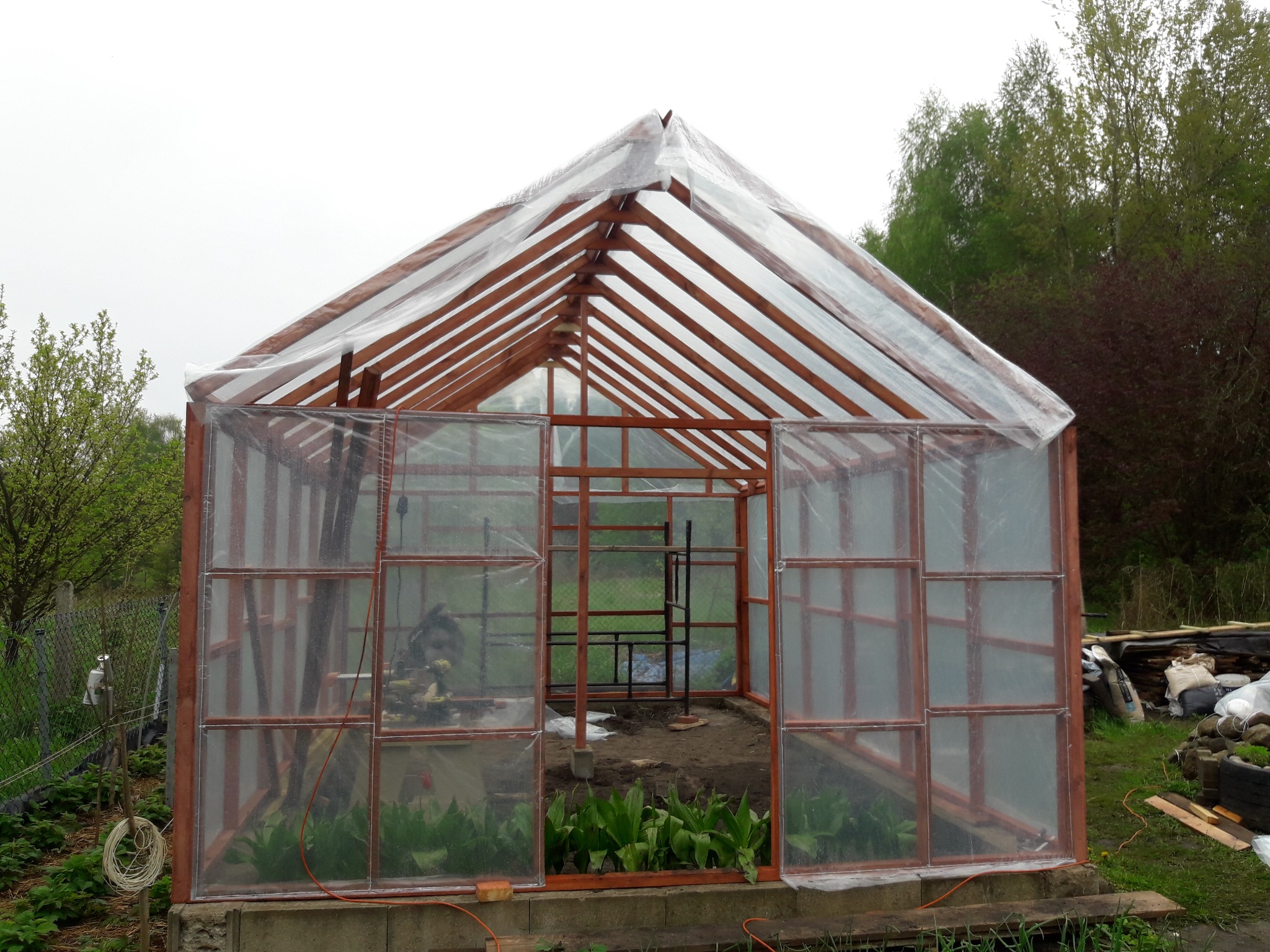 Konstrukcja szklarni z drewnianym szkieletem i przezroczystą folią, widoczne rośliny wewnątrz i narzędzia, otoczenie ogrodowe.