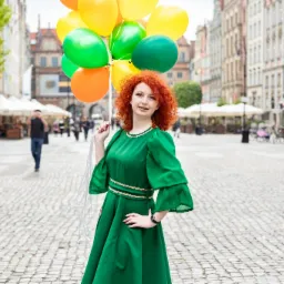 Uśmiechnięta, ruda dziewczyna w zielonej sukience trzyma pęk kolorowych balonów na tle staromiejskiej uliczki z parasolami kawiarnianymi.