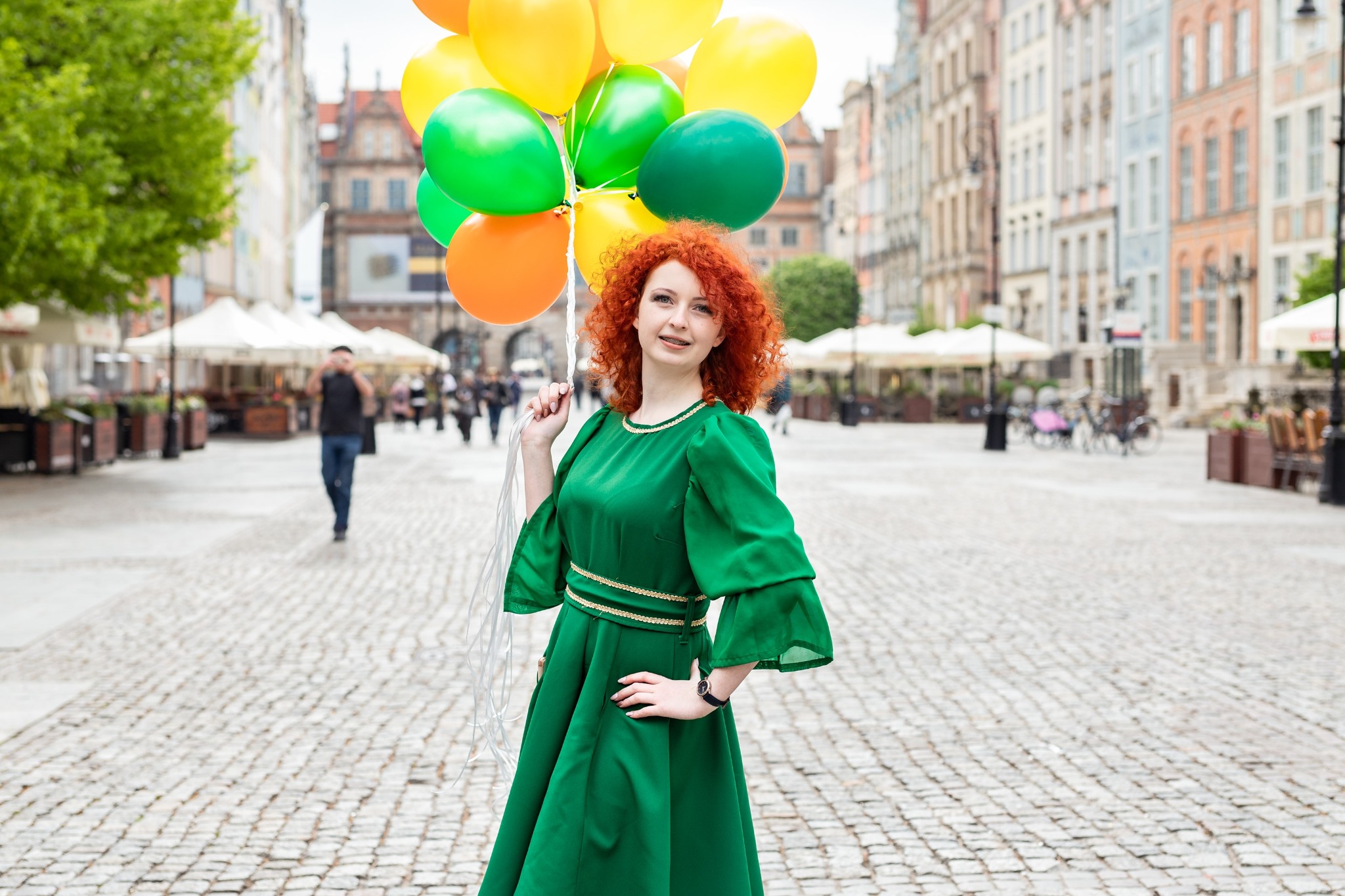 Uśmiechnięta, ruda dziewczyna w zielonej sukience trzyma pęk kolorowych balonów na tle staromiejskiej uliczki z parasolami kawiarnianymi.