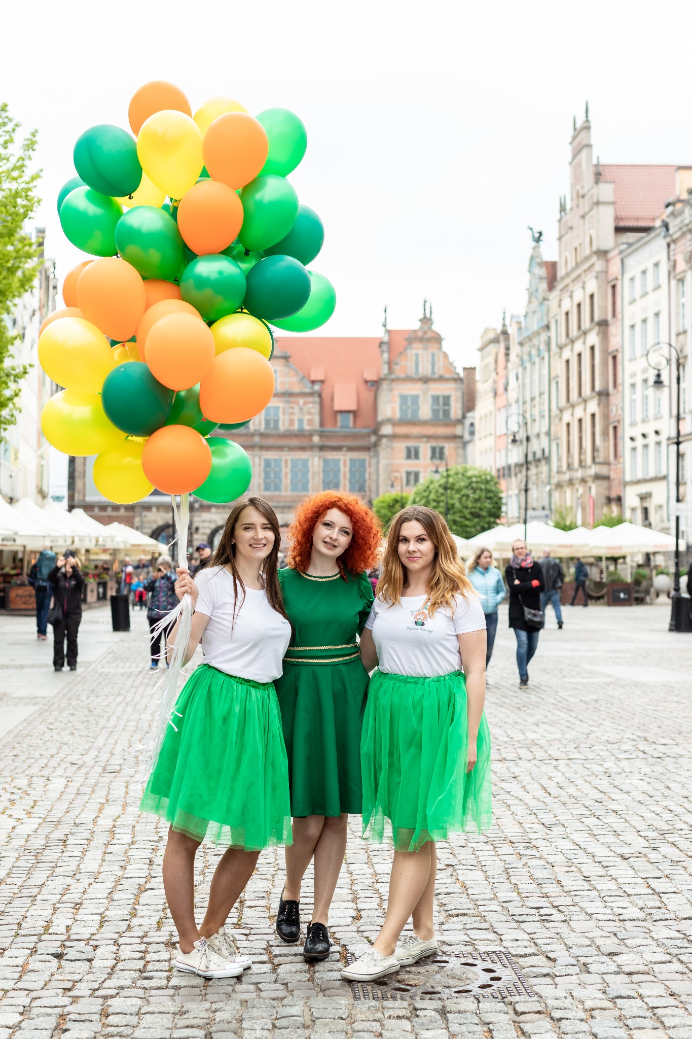 Trzy uśmiechnięte kobiety w zielonych strojach (spódnice tiulowe, sukienka) trzymające bukiet zielono-żółto-pomarańczowych balonów na tle starówki.