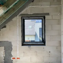 Wnętrze w stanie surowym z widocznymi przewodami elektrycznymi poprowadzonymi po ścianie z betonu komórkowego, okno z widokiem na dom z panelami słonecznymi.