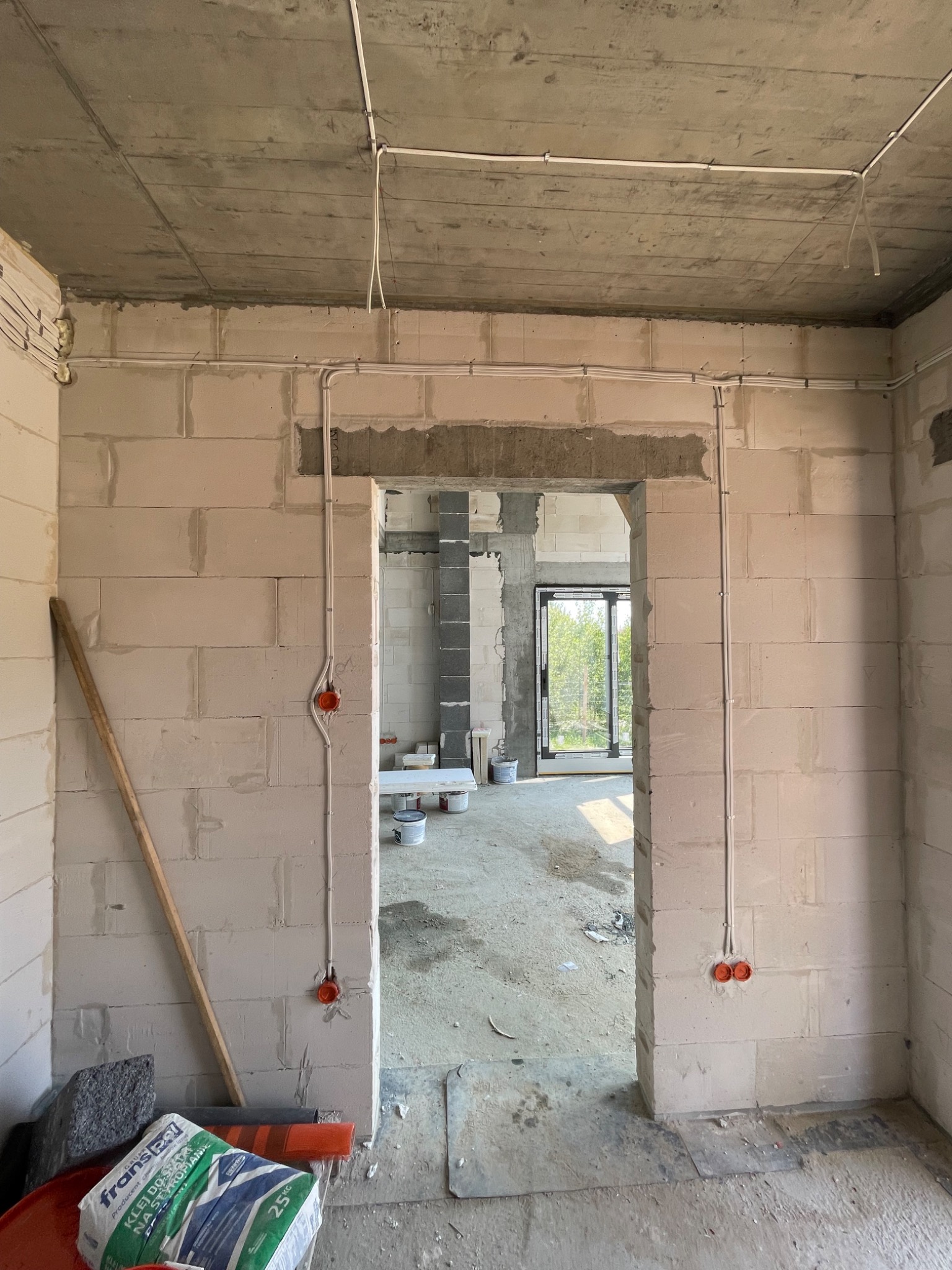 Wnętrze w stanie surowym z widocznymi przewodami elektrycznymi poprowadzonymi po ścianach z betonu komórkowego i pod sufitem, z zamontowanymi puszkami instalacyjnymi w kolorze pomarańczowym.