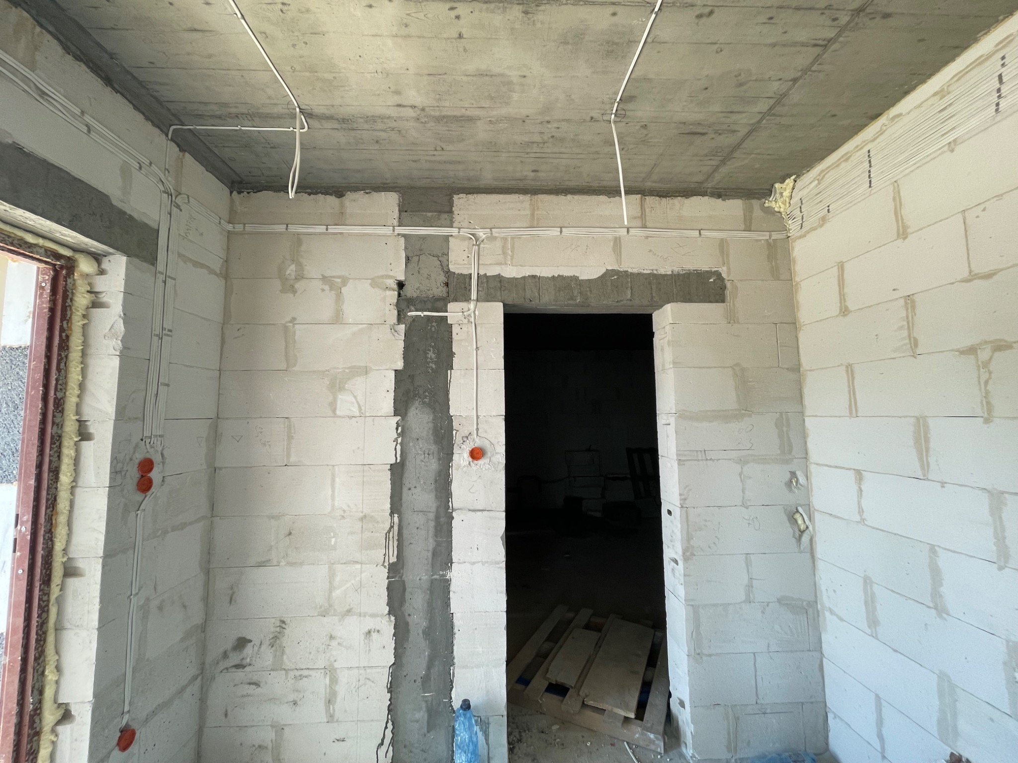 Surowy stan wnętrza z widocznymi przewodami elektrycznymi biegnącymi po suficie i ścianach z betonu komórkowego, pomarańczowe puszki instalacyjne, widoczne ślady zaprawy murarskiej.