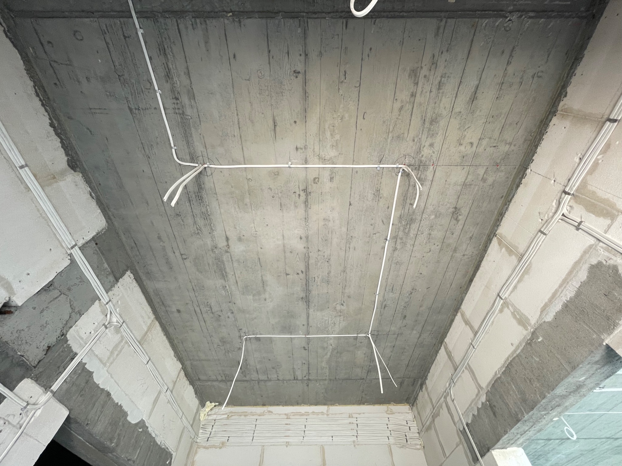 Surowy betonowy sufit z widocznym układem przewodów elektrycznych, zamocowanych do sufitu i ścian w trakcie instalacji, widoczne bloczki betonowe i pianka montażowa.