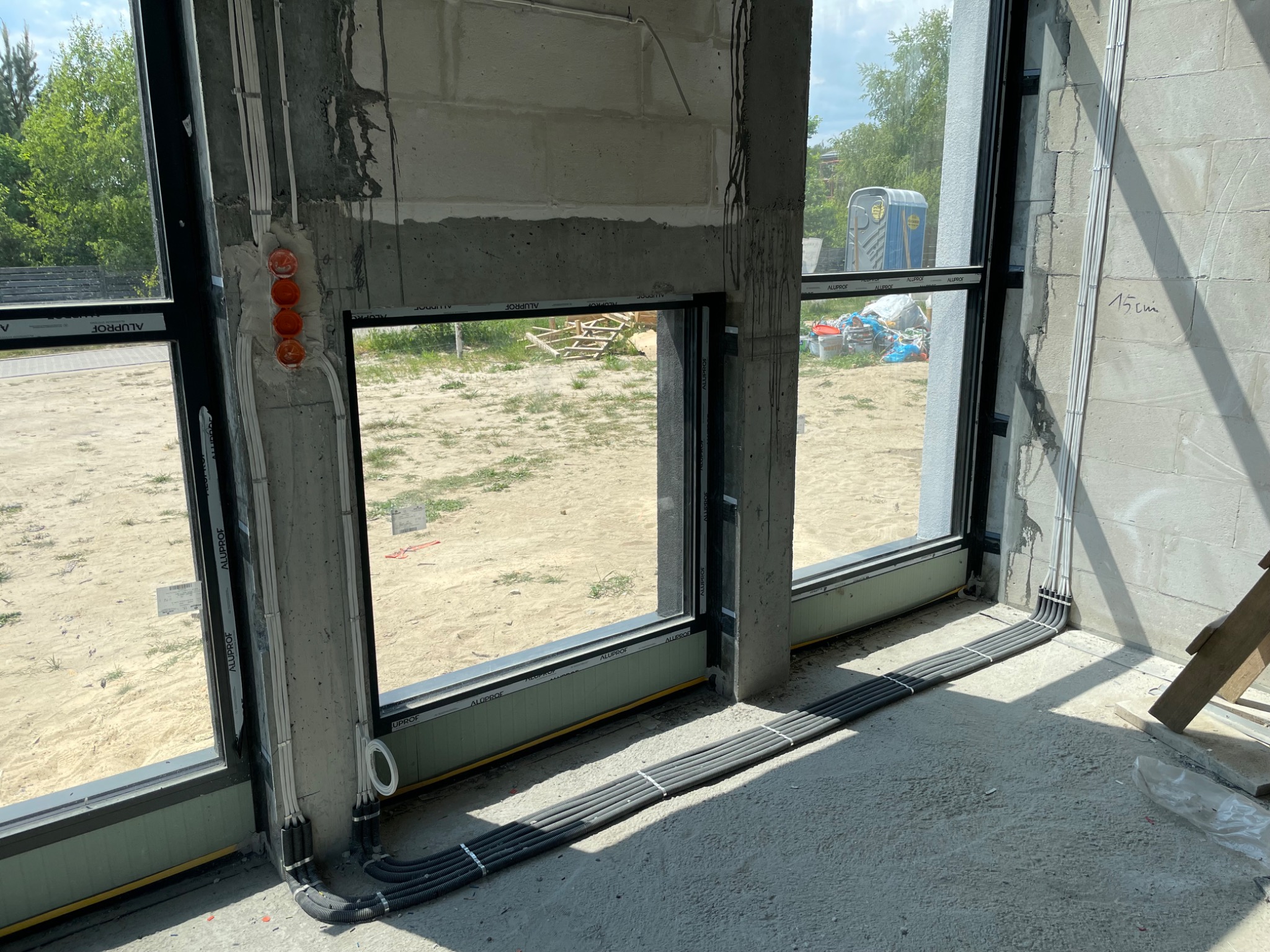 Wnętrze w surowym stanie z widocznymi przewodami elektrycznymi w szarych peszlach ułożonymi wzdłuż ściany i okien, puszki instalacyjne pomarańczowe, widok na zewnątrz przez okno z widoczną budową...