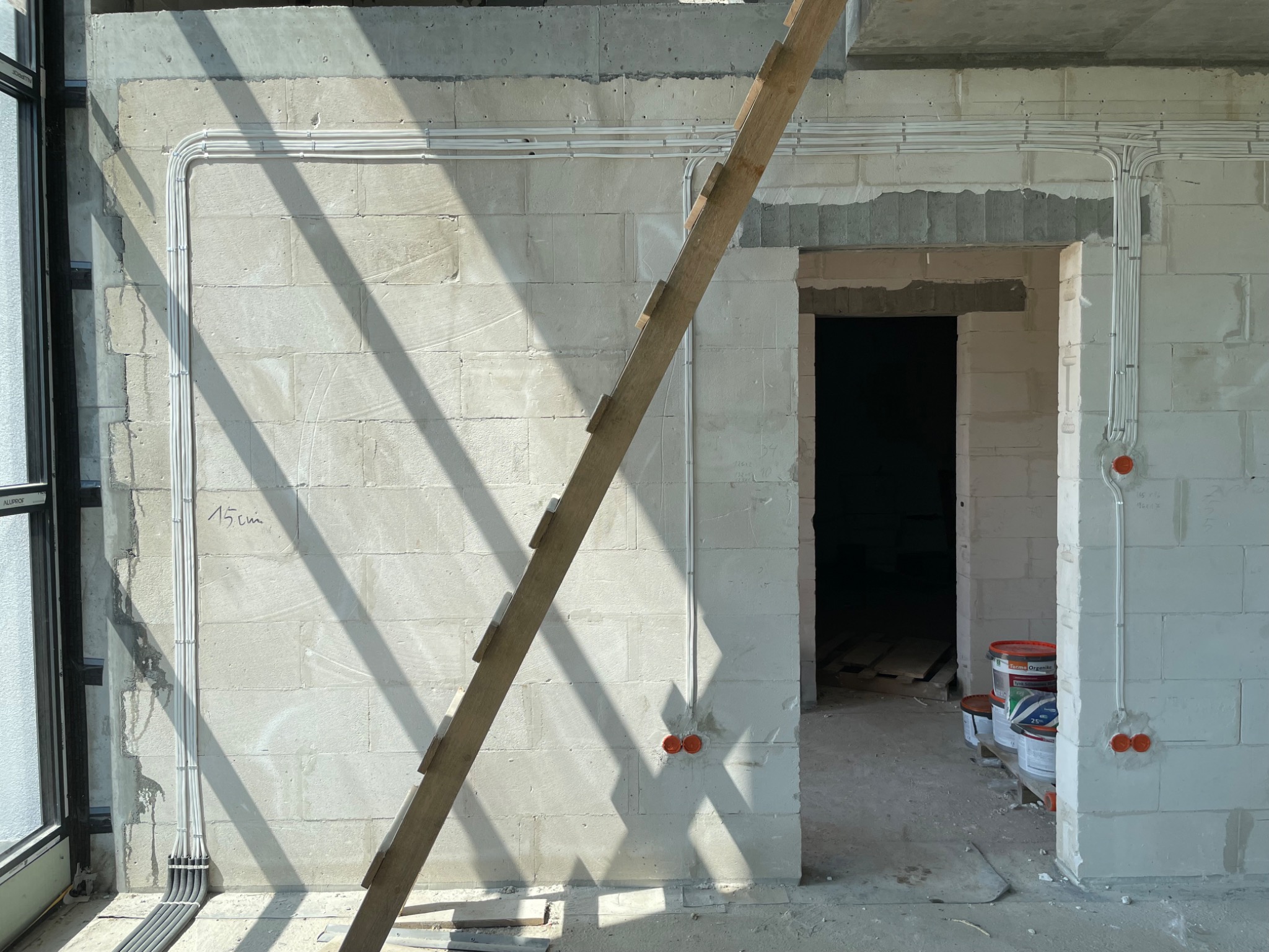 Świeżo położone przewody elektryczne w białych rurkach na ścianie z betonu komórkowego, widoczne puszki instalacyjne i drewniana drabina oparta o ścianę, w tle widoczne puszki z farbą i otwarty...