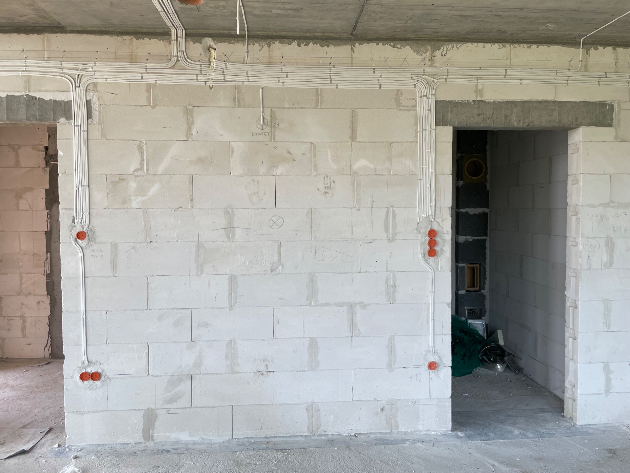 Surowa ściana z betonu komórkowego w trakcie instalacji elektrycznej: widoczne kable w białych rurkach PCV, pomarańczowe puszki pod gniazdka i włączniki, napisy techniczne na ścianie, przygotowane...