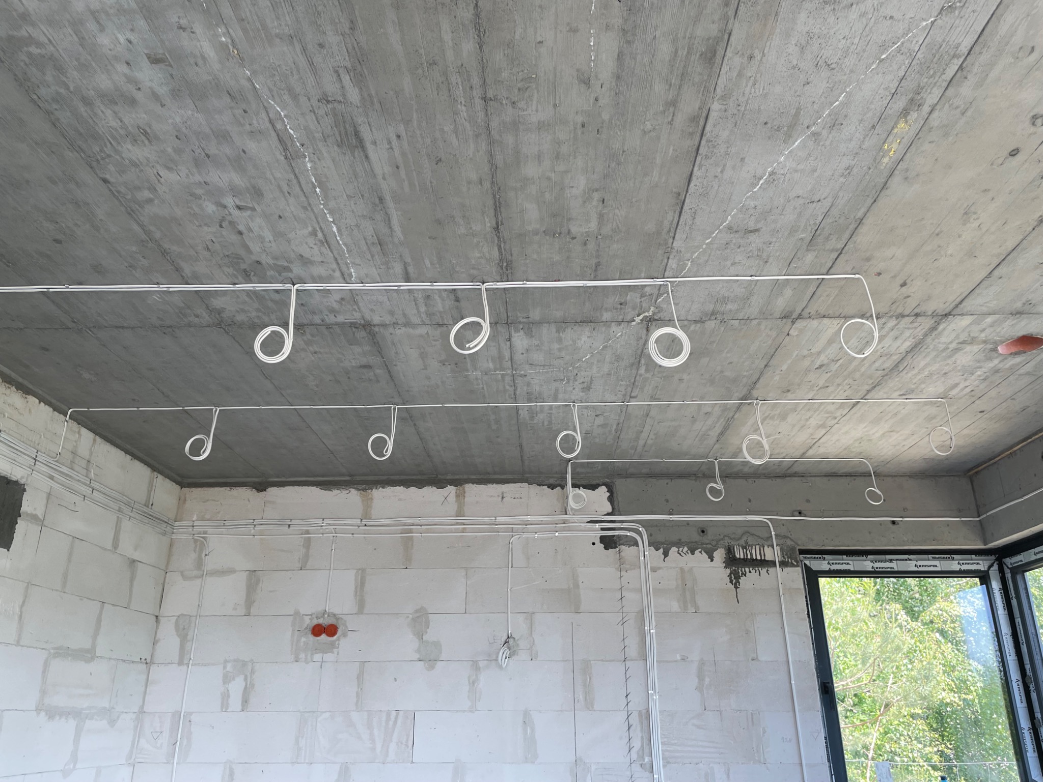 Surowy betonowy sufit z zamocowanymi białymi kablami elektrycznymi, zwisającymi w pętlach, ściany z białych bloczków, widok z okna na zieleń, instalacja w trakcie.