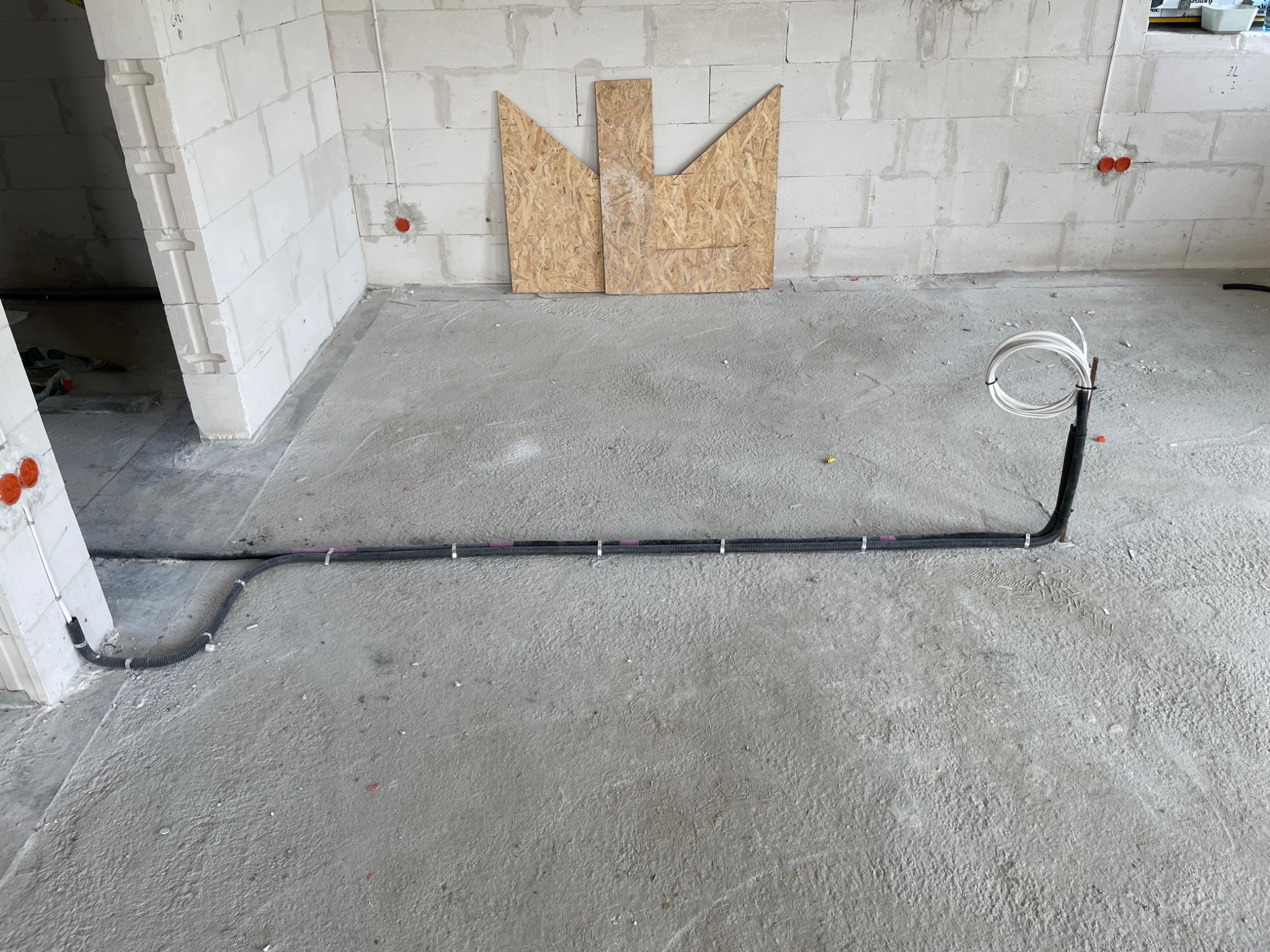 Surowy stan instalacji: czarna karbowana rura z przewodami elektrycznymi ułożona na szarym, betonowym podłożu w pomieszczeniu z białymi ścianami z betonu komórkowego, widoczne puszki instalacyjne...