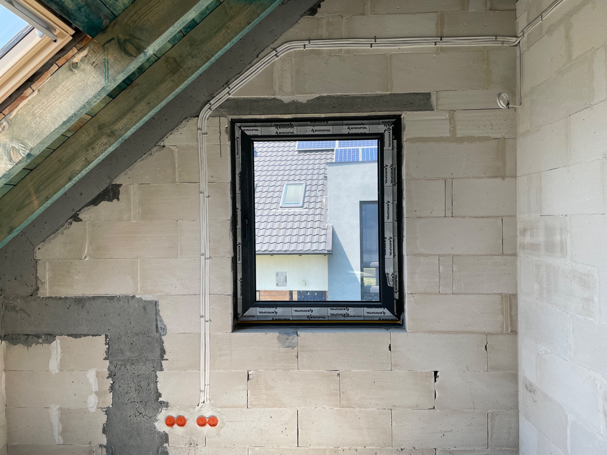 Wnętrze w stanie surowym z widocznymi przewodami elektrycznymi poprowadzonymi po ścianie z betonu komórkowego, okno z widokiem na dom z panelami słonecznymi.