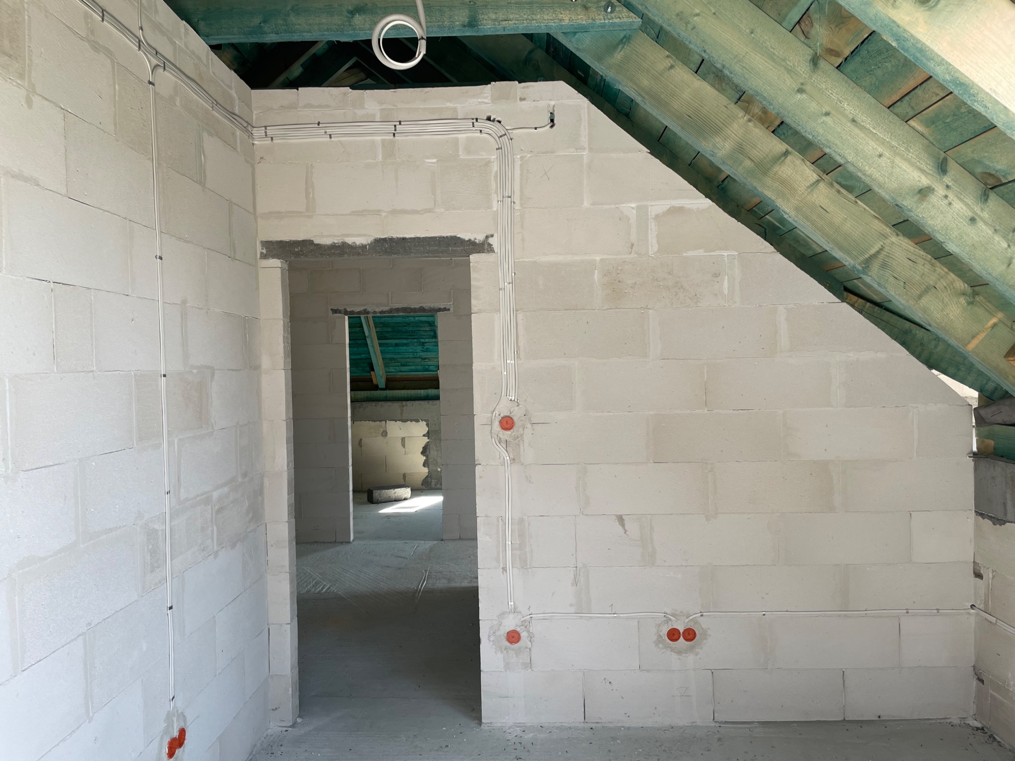 Surowy stan poddasza z białych bloczków, widoczne kable elektryczne poprowadzone po ścianach, puszki instalacyjne w kolorze pomarańczowym, drewniana konstrukcja dachu z zielonymi belkami.