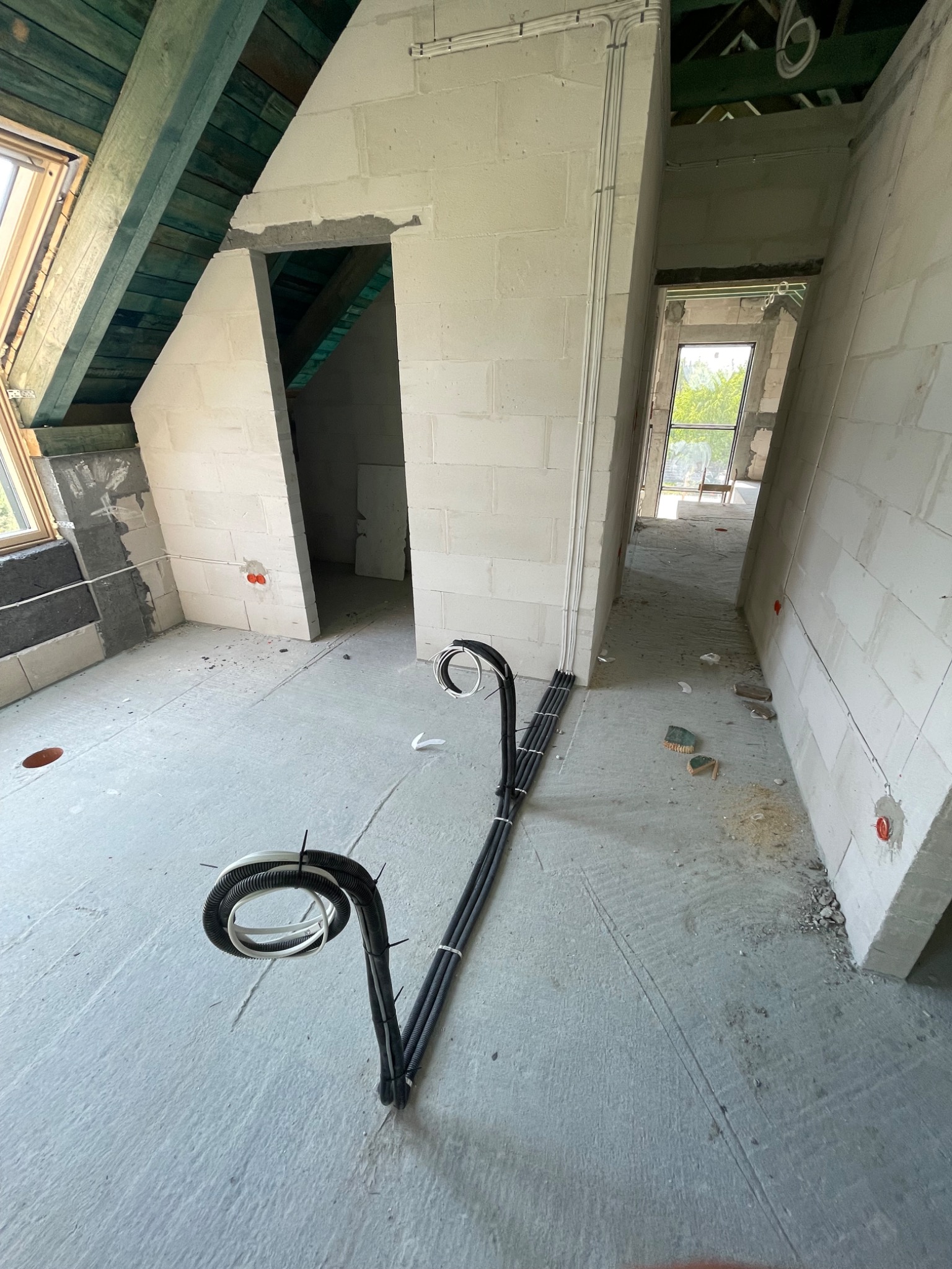 Surowy stan poddasza z widocznymi przewodami elektrycznymi w peszlach, prowadzącymi wzdłuż ściany z betonu komórkowego, z widocznymi otworami w ścianach na gniazdka.