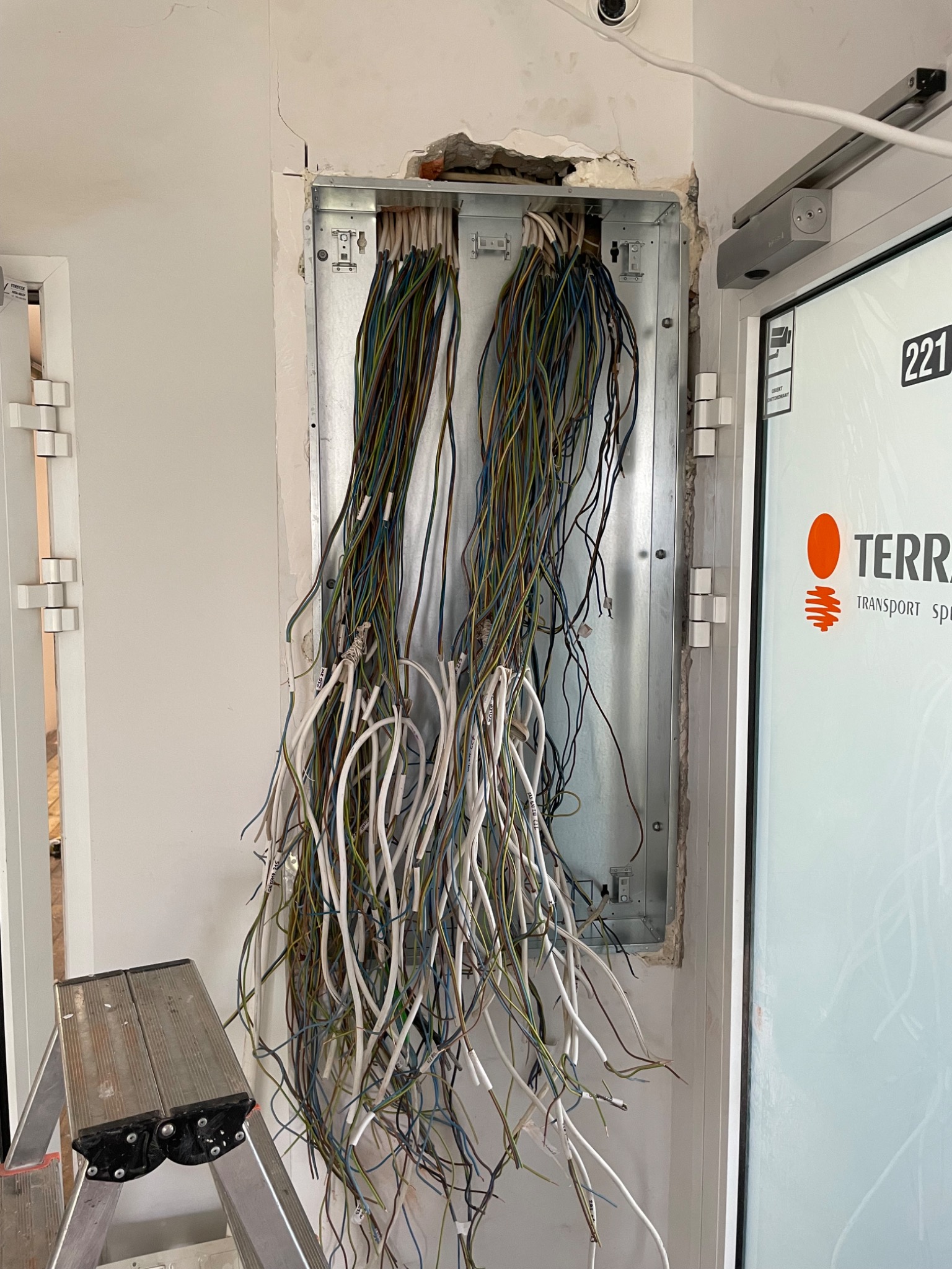 Otwarta skrzynka elektryczna w ścianie z wystającymi kablami o różnych kolorach, widoczna drabina i fragment drzwi z logo firmy.