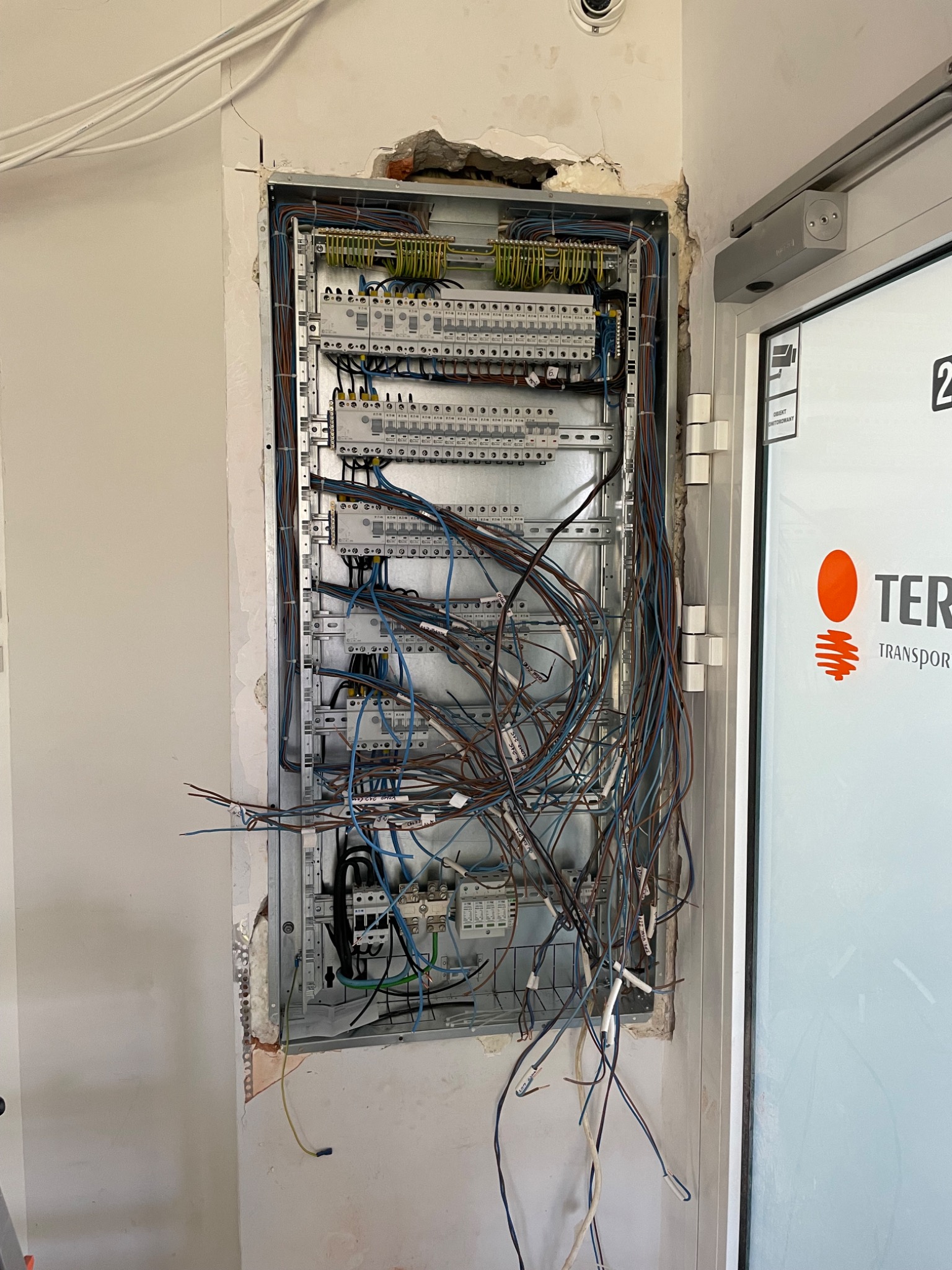 Otwarta skrzynka elektryczna w ścianie z widocznymi przewodami i elementami instalacji, obok drzwi z widocznym logo firmy.