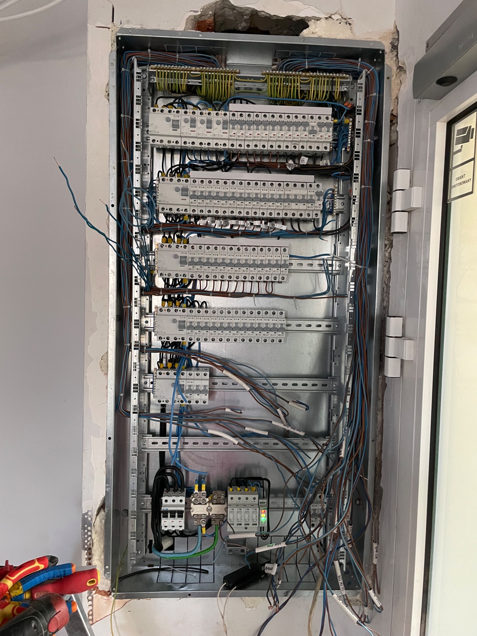 Rozdzielnica elektryczna w trakcie montażu, widoczne liczne przewody, zabezpieczenia i listwy zaciskowe, ściana w stanie surowym, widoczne narzędzia elektryczne.