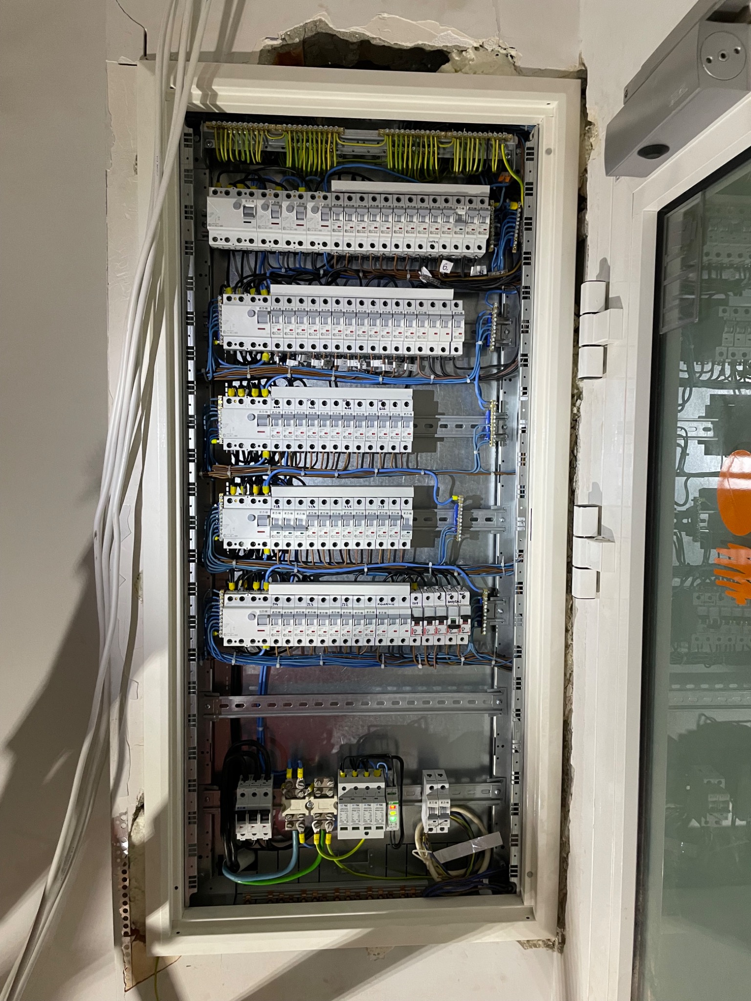 Otwarta skrzynka elektryczna z bezpiecznikami i okablowaniem, zamontowana w ścianie z widocznymi uszkodzeniami tynku.