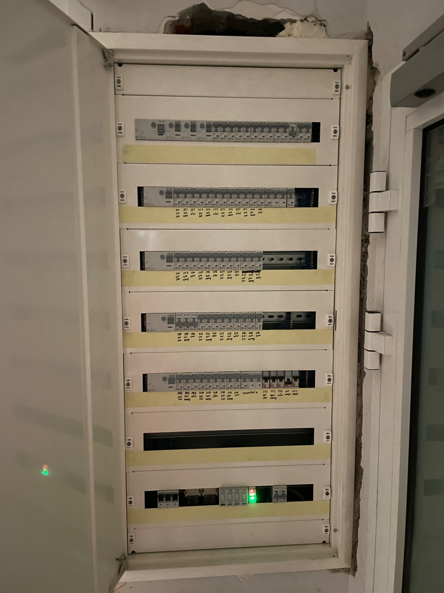 Otwarta skrzynka elektryczna z bezpiecznikami i oznaczeniami, widoczne przewody i elementy instalacji, stan po montażu, ściana z ubytkami tynku.