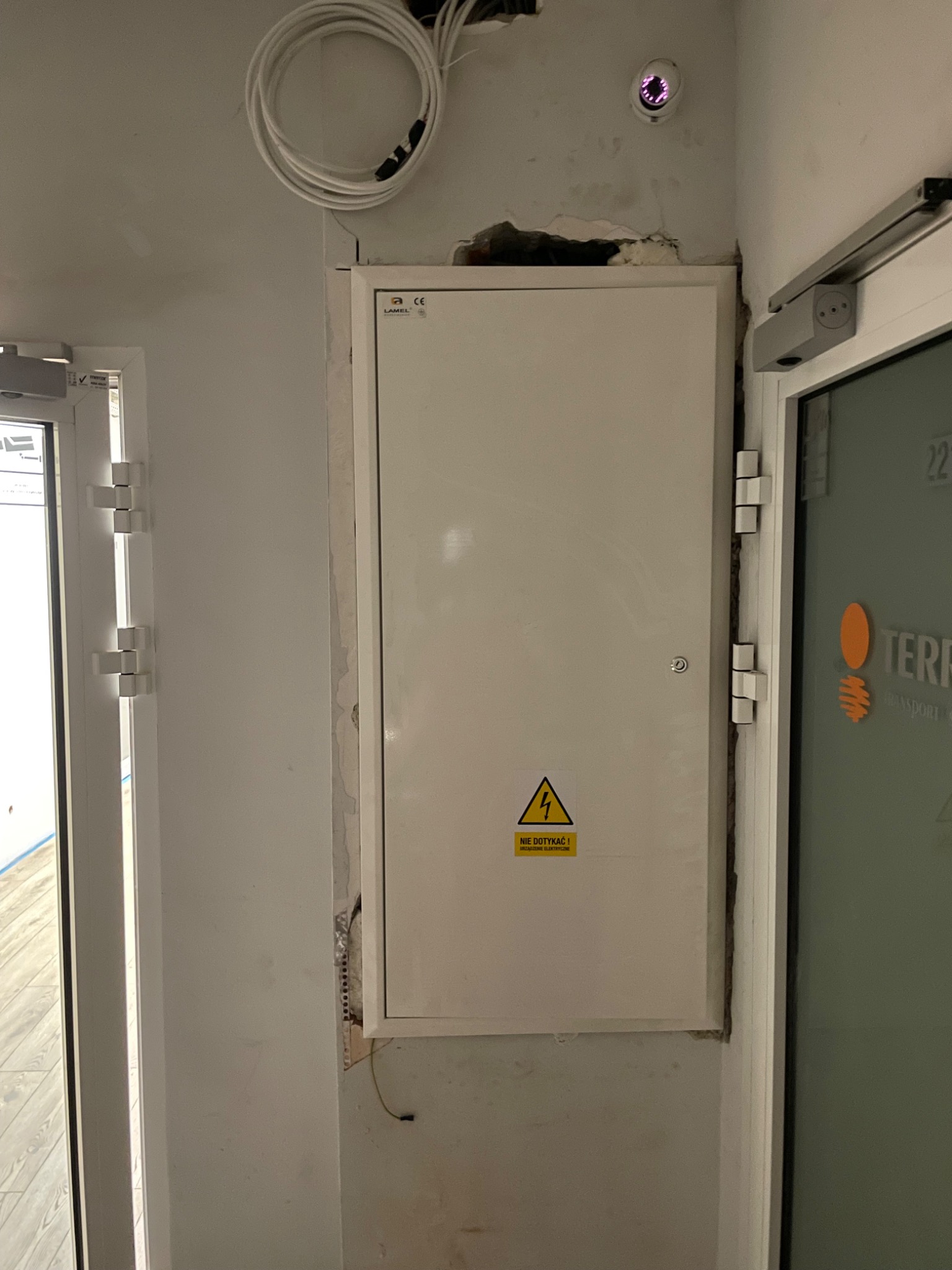 Biała skrzynka elektryczna z ostrzeżeniem 'NIE DOTYKAĆ! Urządzenie elektryczne' na ścianie obok drzwi, widoczne przewody i kamera przemysłowa na suficie.