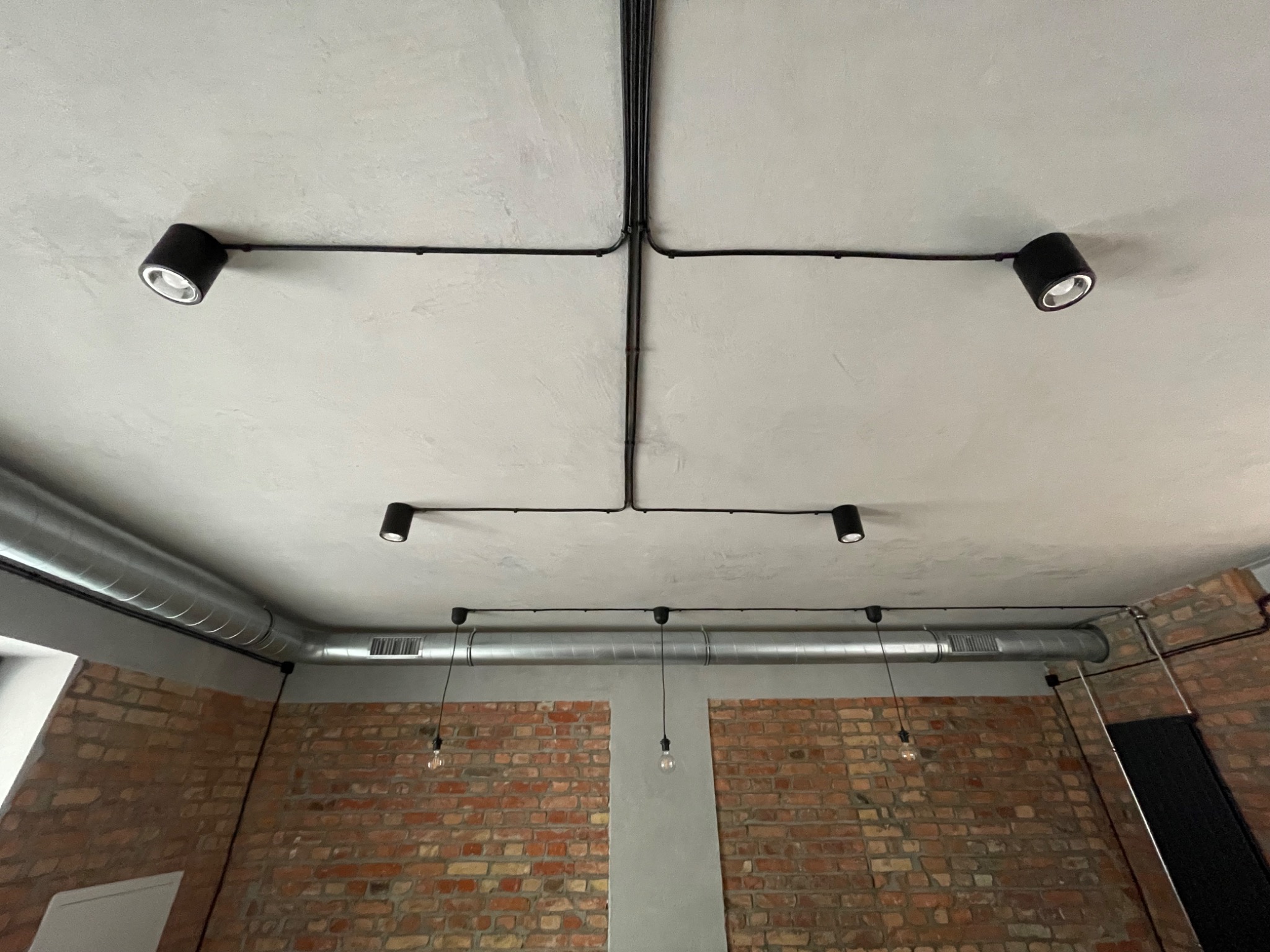 Cztery czarne reflektory punktowe zamontowane na suficie z widocznymi przewodami elektrycznymi, surowy betonowy sufit, wentylacja i ściana z czerwonej cegły w tle.