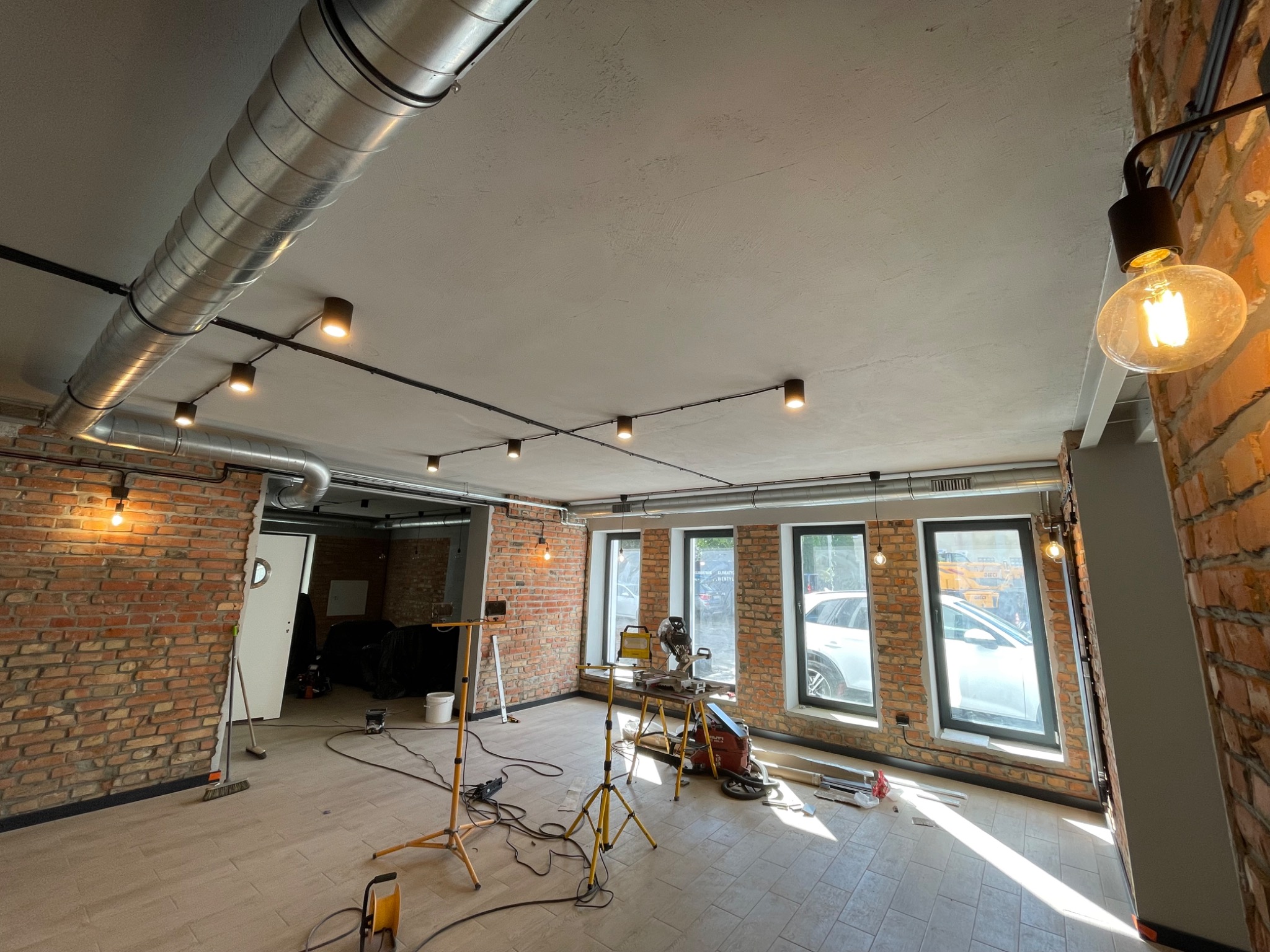 Wnętrze w trakcie remontu z widoczną instalacją oświetleniową: sufitowe reflektory punktowe na szynoprzewodach i wisząca żarówka Edisona na tle ceglanej ściany, narzędzia pracy i kable na podłodze.
