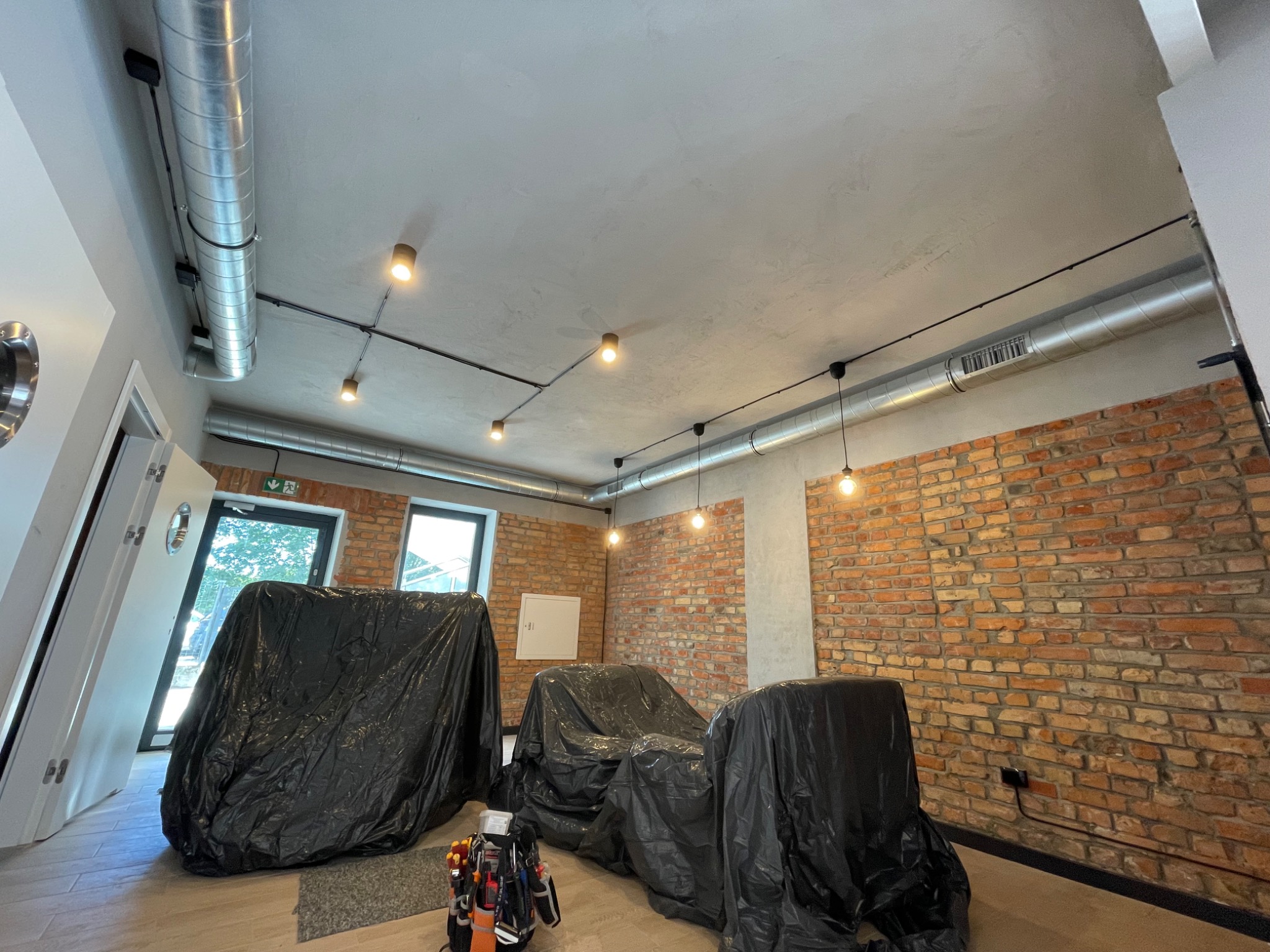 Wnętrze w trakcie montażu oświetlenia: widoczne lampy punktowe na szynoprzewodach, narzędzia elektryczne i meble zabezpieczone folią malarską, ściana z czerwonej cegły.