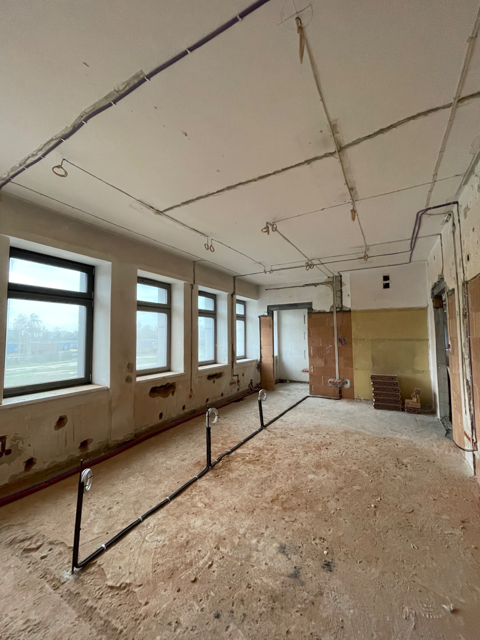 Wnętrze w stanie surowym z widocznymi przewodami elektrycznymi poprowadzonymi po suficie i podłodze, przygotowane do dalszych prac wykończeniowych.