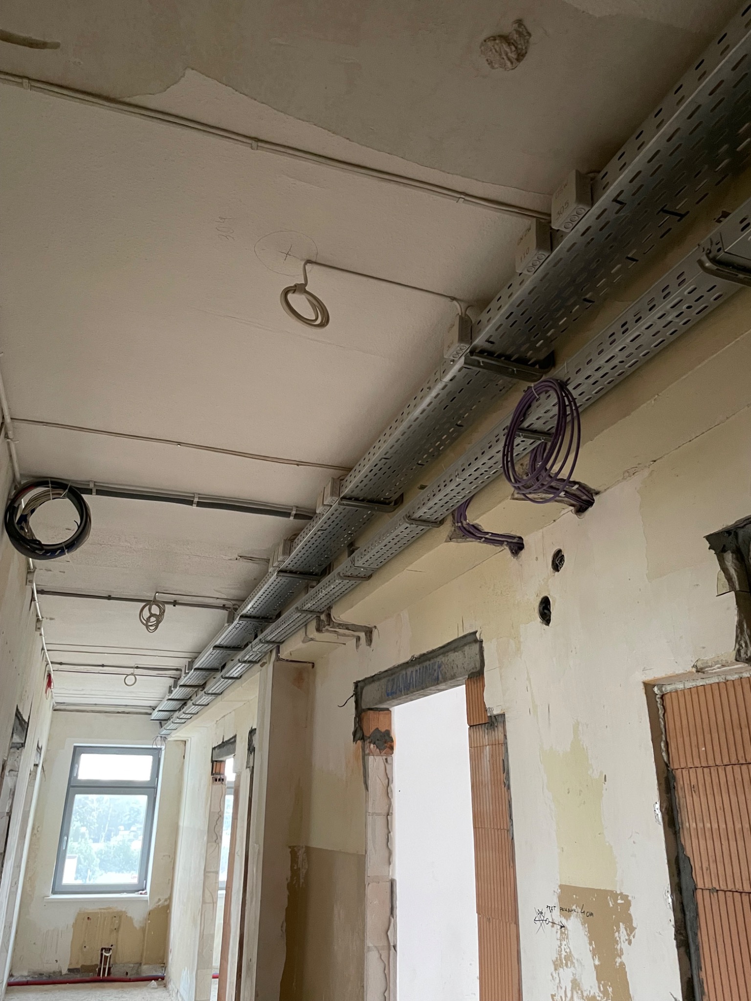 Korytarz w trakcie remontu z widocznymi metalowymi korytkami kablowymi pod sufitem, prowadzącymi przewody elektryczne w kolorze fioletowym, oraz wiszącymi pętlami kabli. Ściany w stanie surowym...