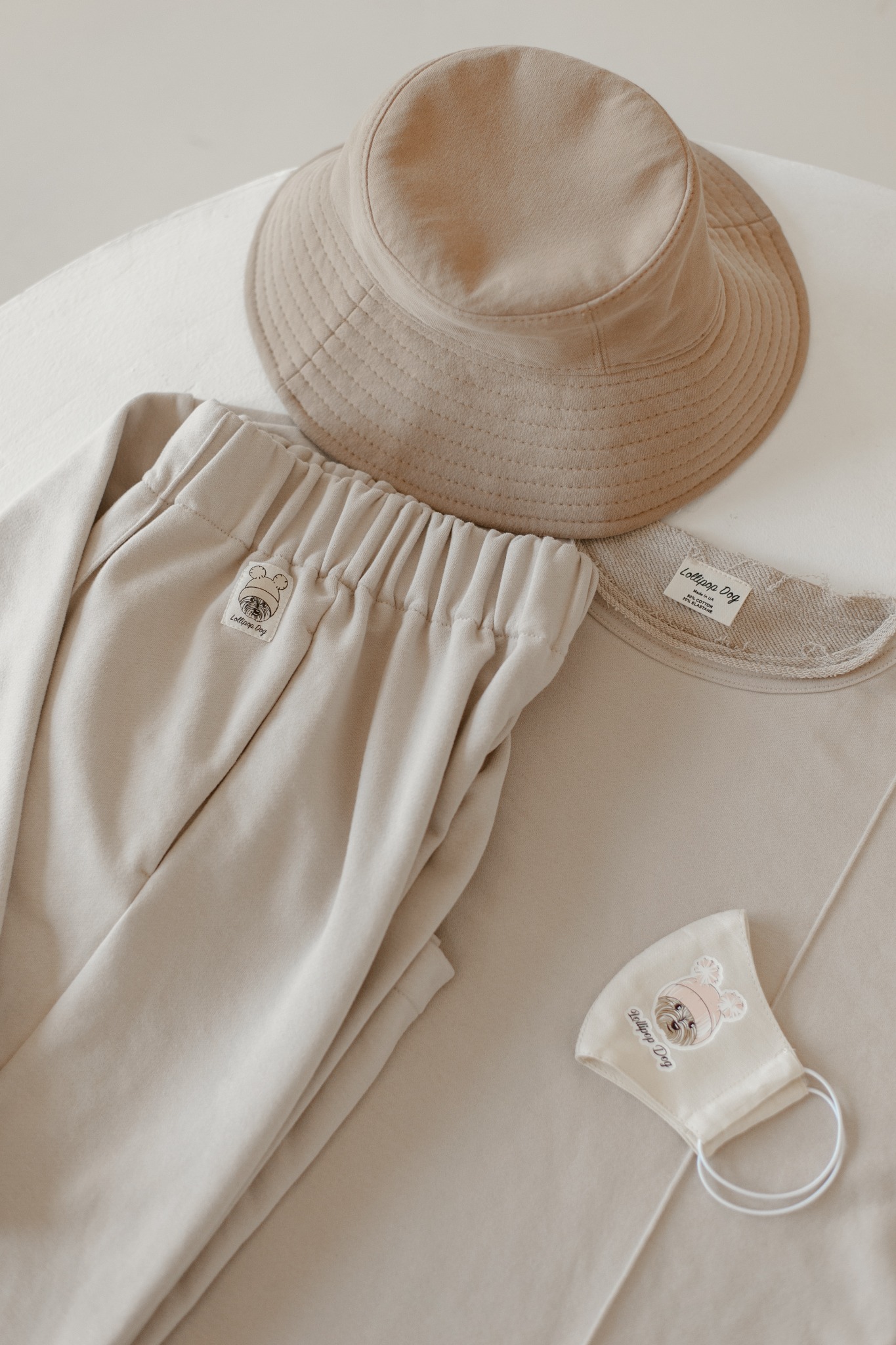 Beżowy komplet odzieżowy: трикотажный спортивный женский костюм składający się z bluzy оверсайз z необработанными краями i szerokich spodni, панама oraz maseczka ochronna z nadrukiem logo firmy.