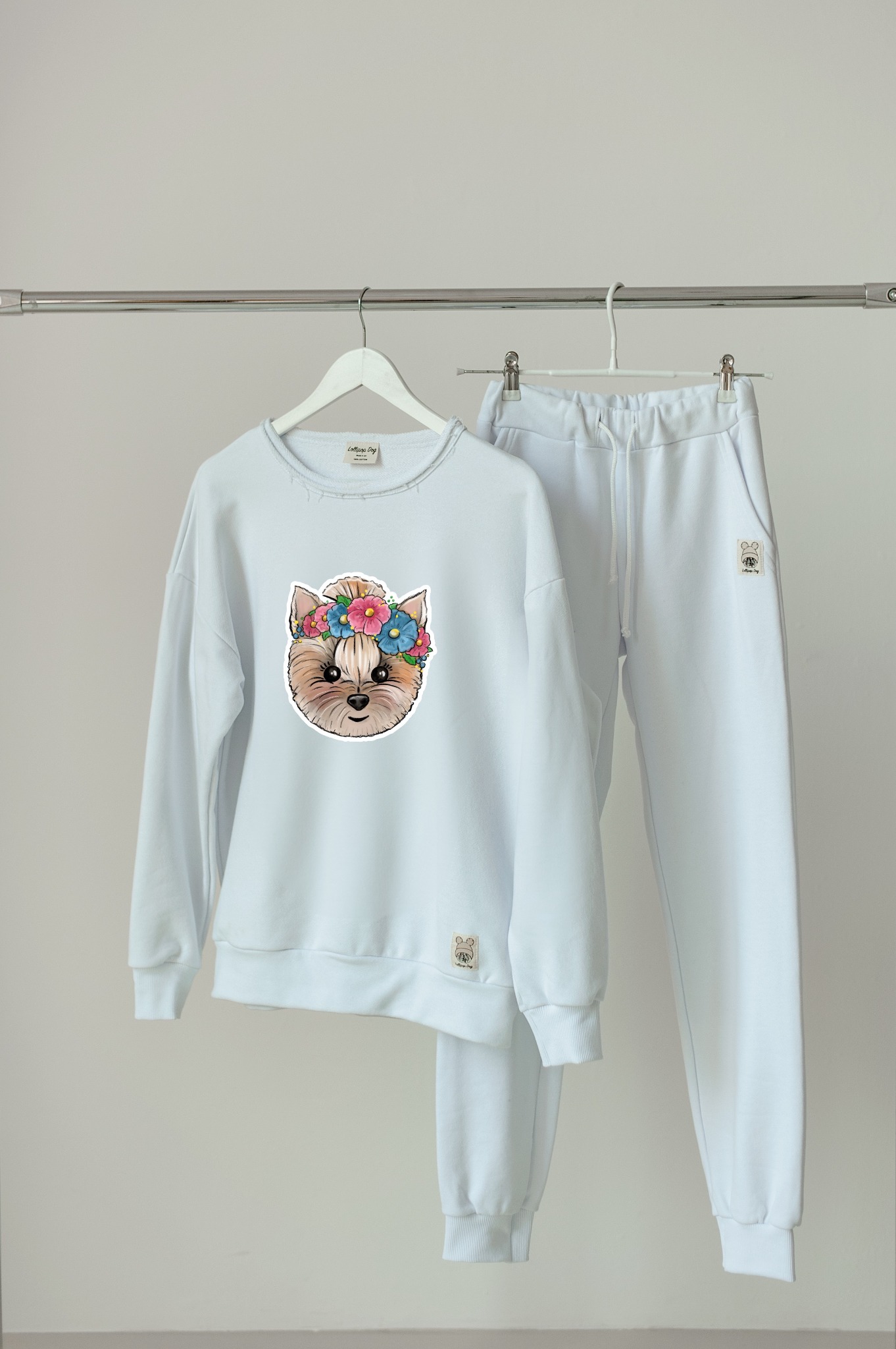 Jasny, damski dres sportowy z nadrukiem głowy psa w wianku z kwiatów, wiszący na wieszaku na tle jasnej ściany.