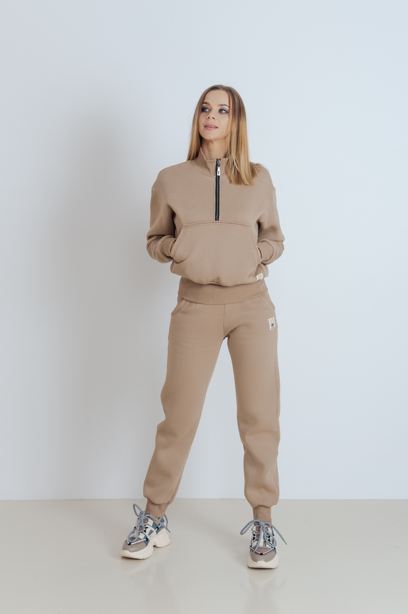 Kobieta prezentuje beżowy dres z dzianiny: bluza z krótkim zamkiem i kieszenią kangurką oraz spodnie typu jogger z wysokim stanem. Całość uzupełniają masywne sportowe buty.