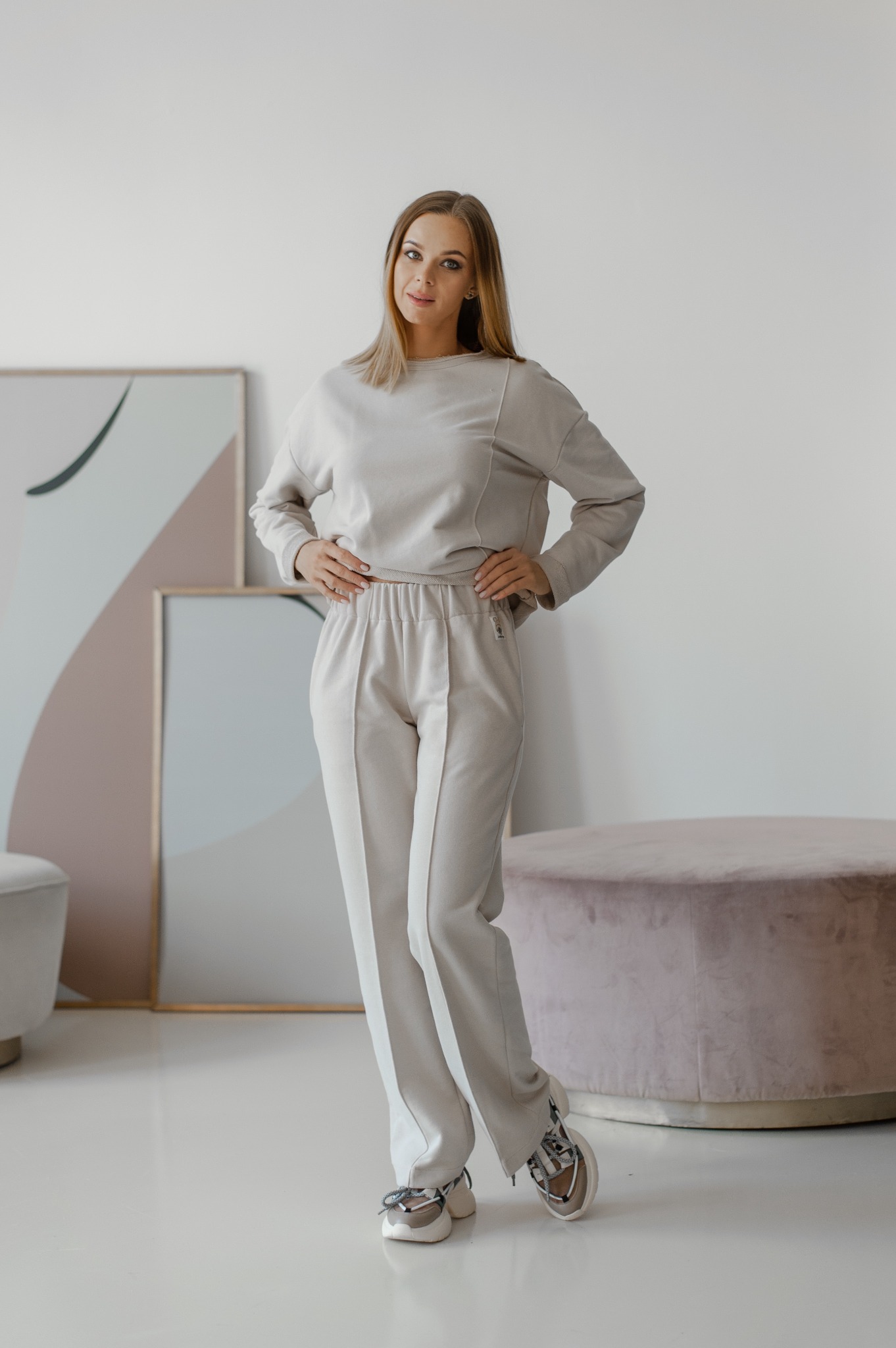 Kobieta prezentuje beżowy, sportowy komplet dresowy (bluza oversize i szerokie spodnie ze strzałkami) w minimalistycznym wnętrzu w Kyiv. Widoczne detale: niewykończone brzegi bluzy, wysoki stan...