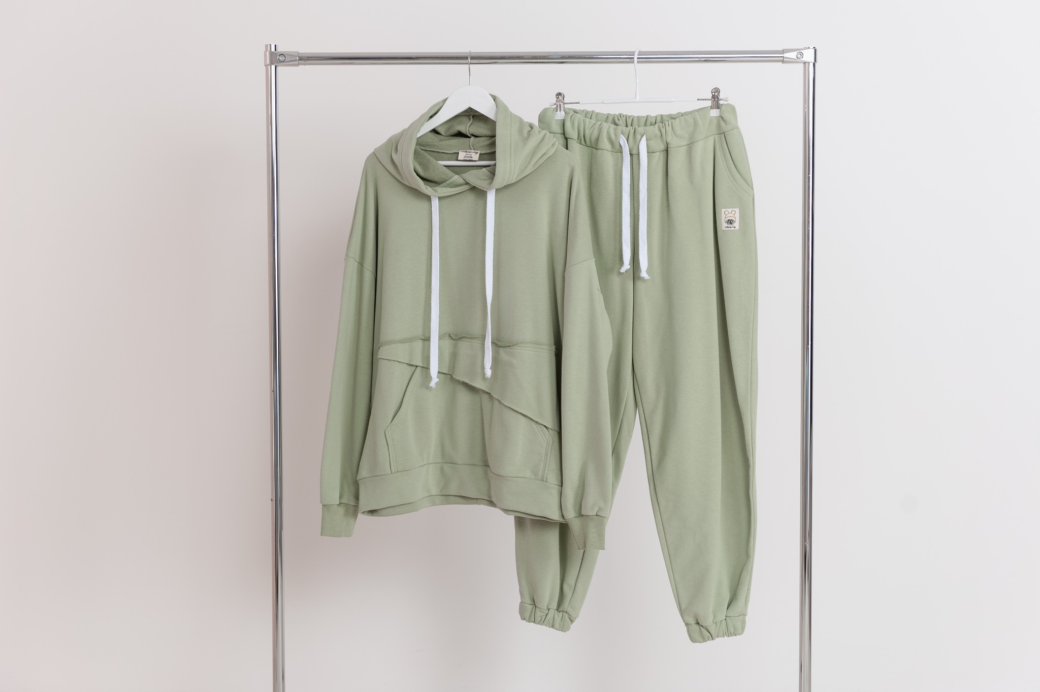 Jasnozielony dres sportowy z трикотажа 3-нить, пенье, składający się z bluzy oversize z kapturem i asymetryczną kieszenią oraz spodni typu jogger z wysokim stanem, wiszący na metalowym wieszaku...