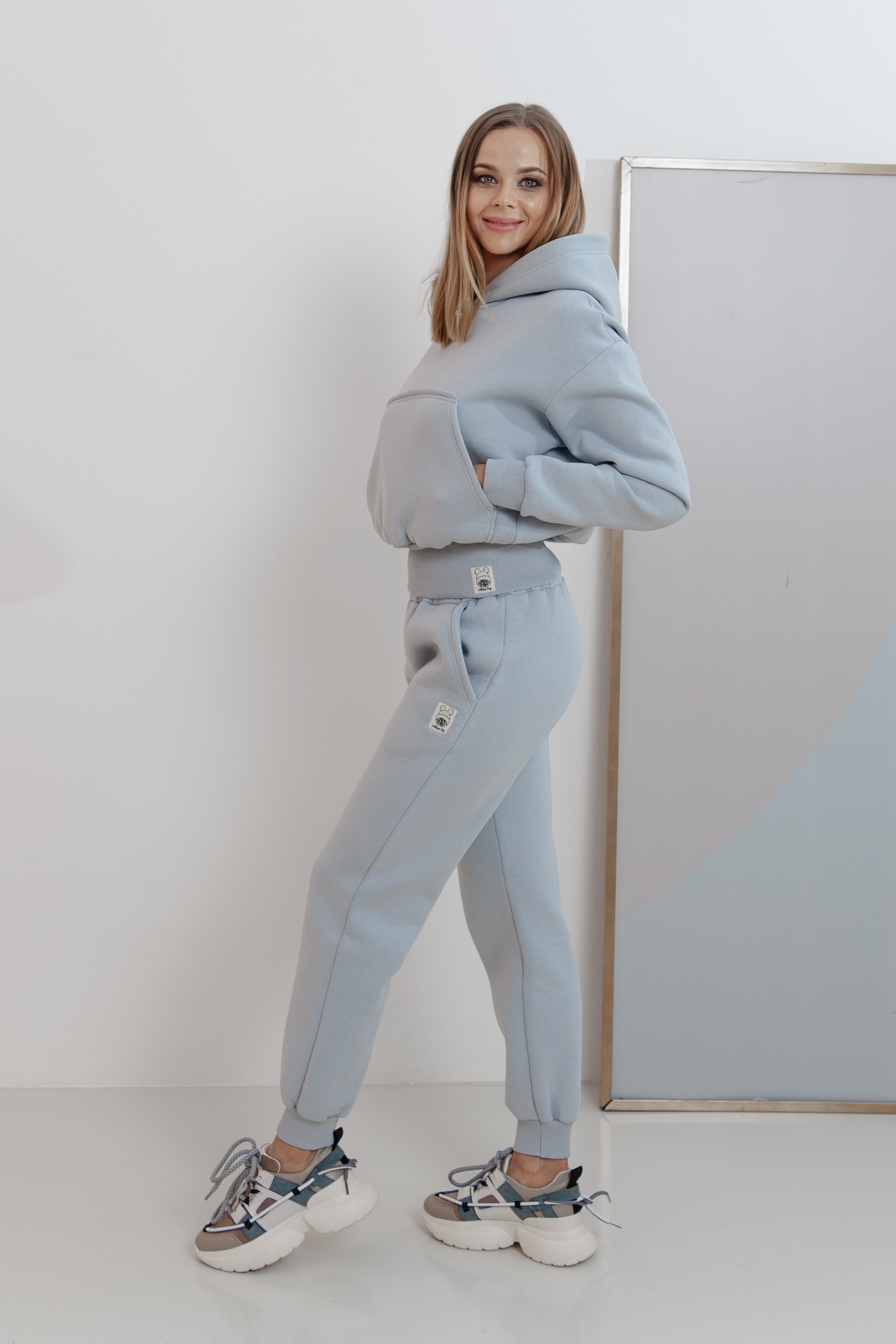 Kobieta w błękitnym, dresowym komplecie z kapturem i spodniami typu jogger, pozująca na jasnym tle z minimalistycznym obrazem. Widoczne masywne sportowe buty.