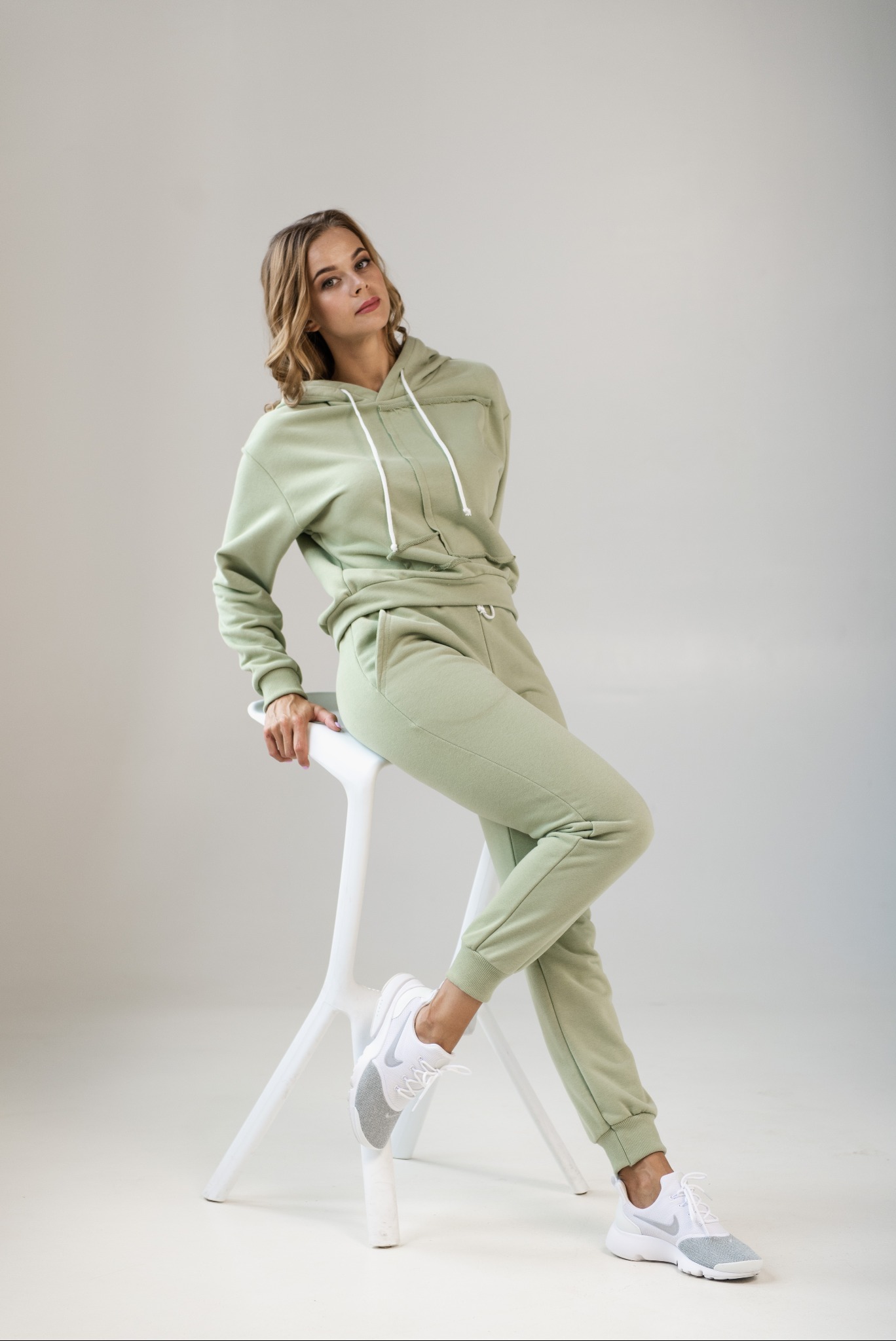 Kobieta pozuje siedząc na białym stołku w pistacjowym, dresowym komplecie z kapturem i białych sportowych butach na neutralnym tle.