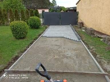 Etap prac przy układaniu szarej kostki brukowej na przygotowanym podłożu, widoczne obrzeża z kostki, ubijarka w tle, tło z zieleni, brama wjazdowa i budynek.