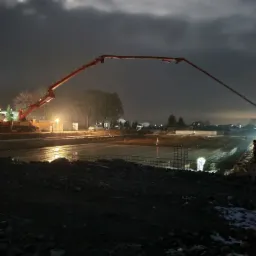 Nocne wylewanie betonu z użyciem pompy, widoczne zbrojenie i oświetlenie budowlane, w tle zabudowania.