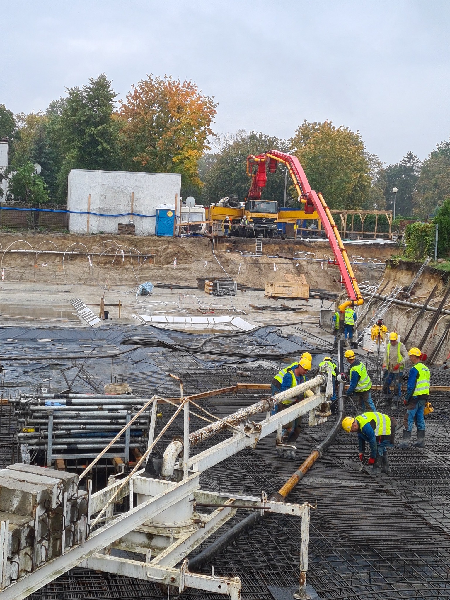 Prace wylewkowe na budowie: ekipa w żółtych kamizelkach rozprowadza beton z pompy na zbrojeniu, w tle wykop i drzewa w jesiennych barwach.