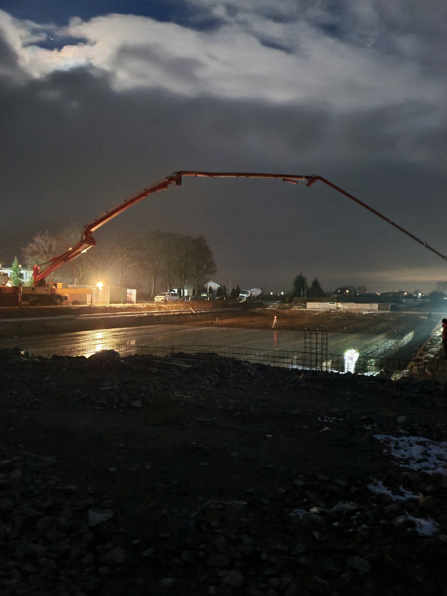 Nocne wylewanie betonu z użyciem pompy, widoczne zbrojenie i oświetlenie budowlane, w tle zabudowania.