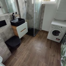 Fraktal - Nowoczesna łazienka z brązowymi płytkami podłogowymi imitującymi drewno, czarną umywalką nablatową na szafce, kabiną prysznicową z czarnymi profilami, toaletą w kolorze czarnym oraz pralką.