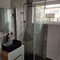 Nowoczesna łazienka z czarną umywalką nablatową, kabiną prysznicową typu walk-in i ścianami wyłożonymi patchworkowymi płytkami w odcieniach szarości, widok na budynek z okna.