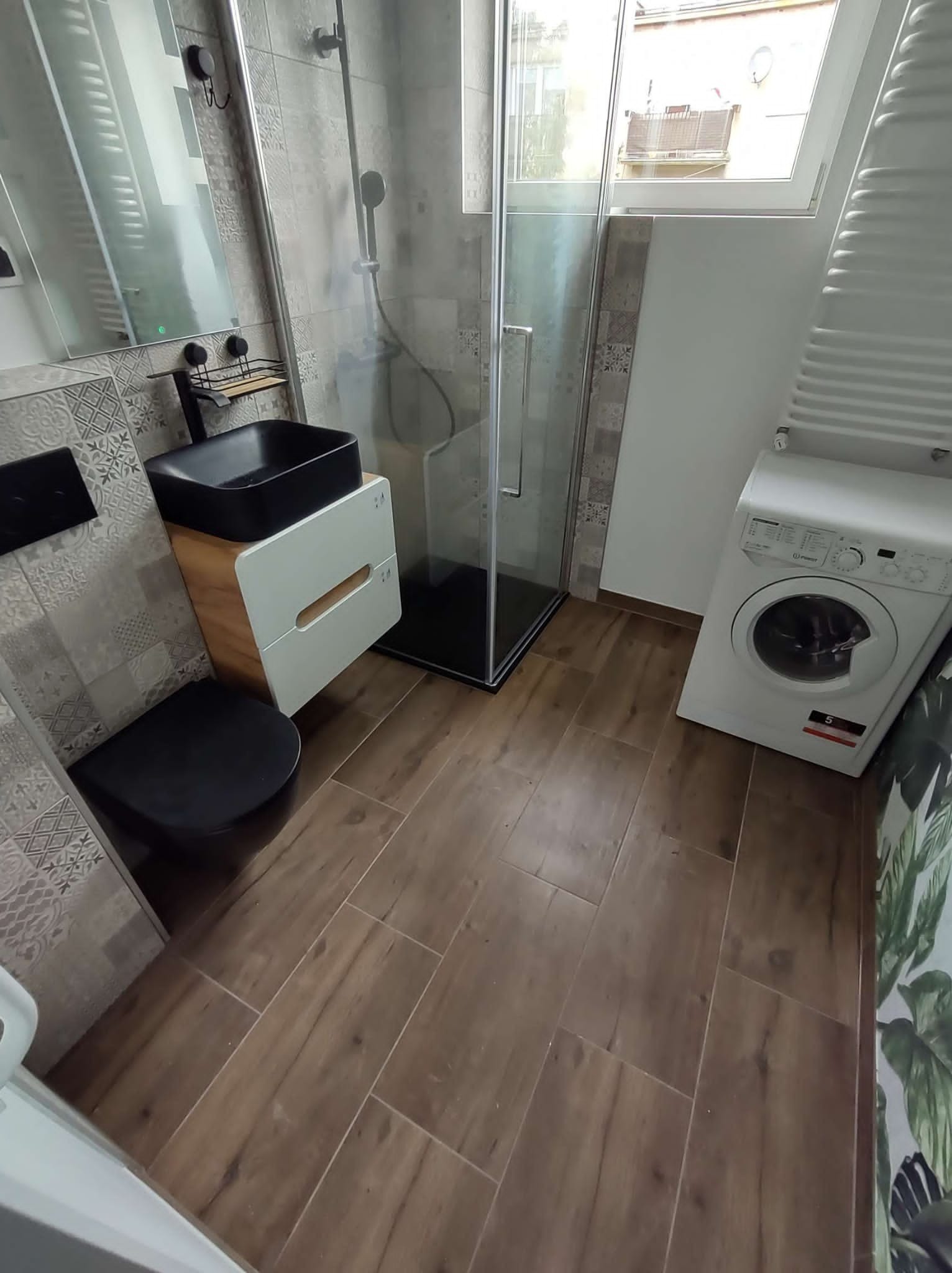 Nowoczesna łazienka z brązowymi płytkami podłogowymi imitującymi drewno, czarną umywalką nablatową na szafce, kabiną prysznicową z czarnymi profilami, toaletą w kolorze czarnym oraz pralką.