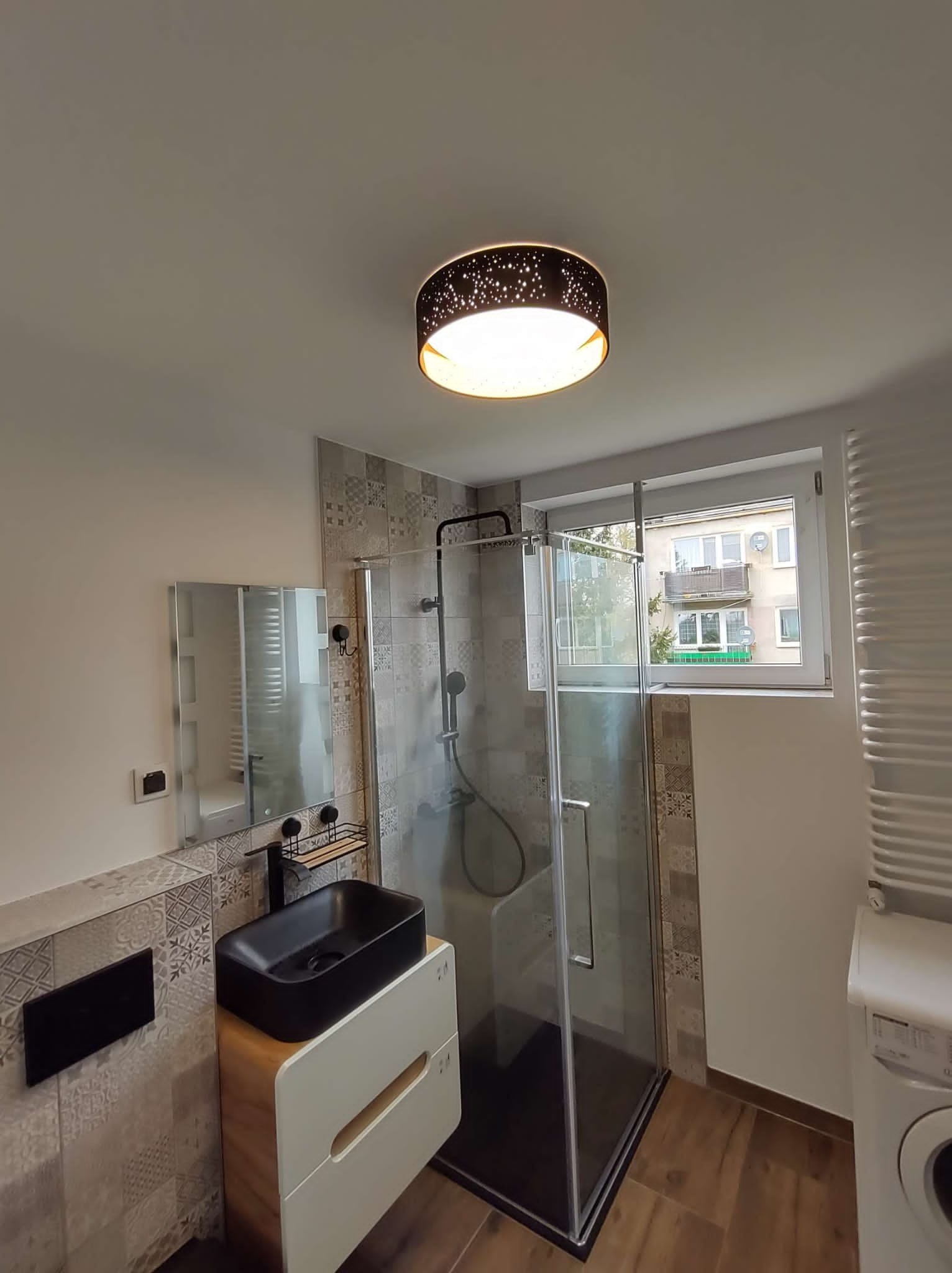 Nowoczesna łazienka po remoncie: kabina prysznicowa typu walk-in z czarną armaturą, umywalka nablatowa w kolorze czarnym, podwieszana szafka z drewnianymi akcentami, dekoracyjne płytki na ścianach,...