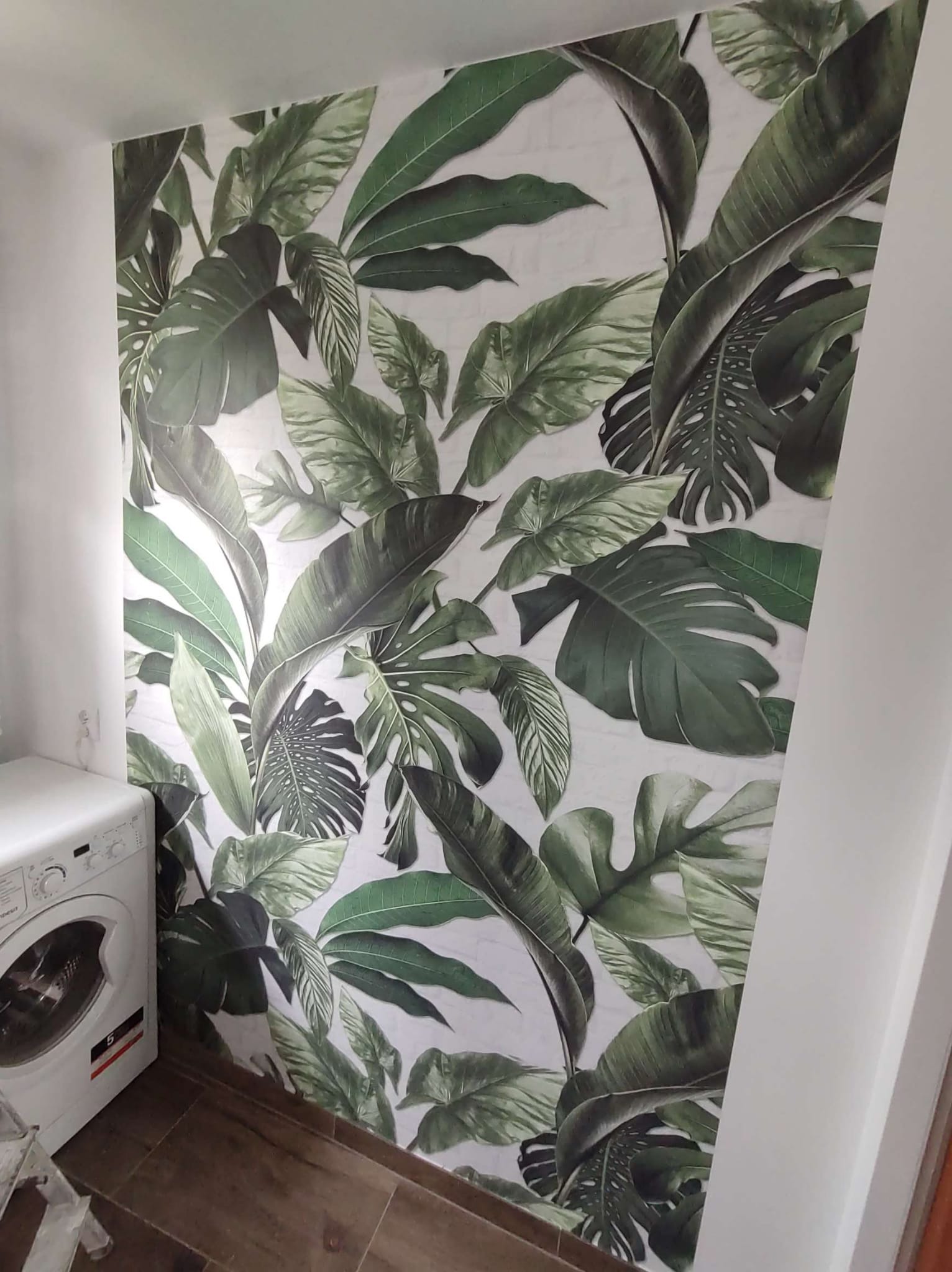 Ściana w Krakowie wyłożona płytkami z motywem tropikalnych liści w łazience obok pralki, z drewnopodobną podłogą.