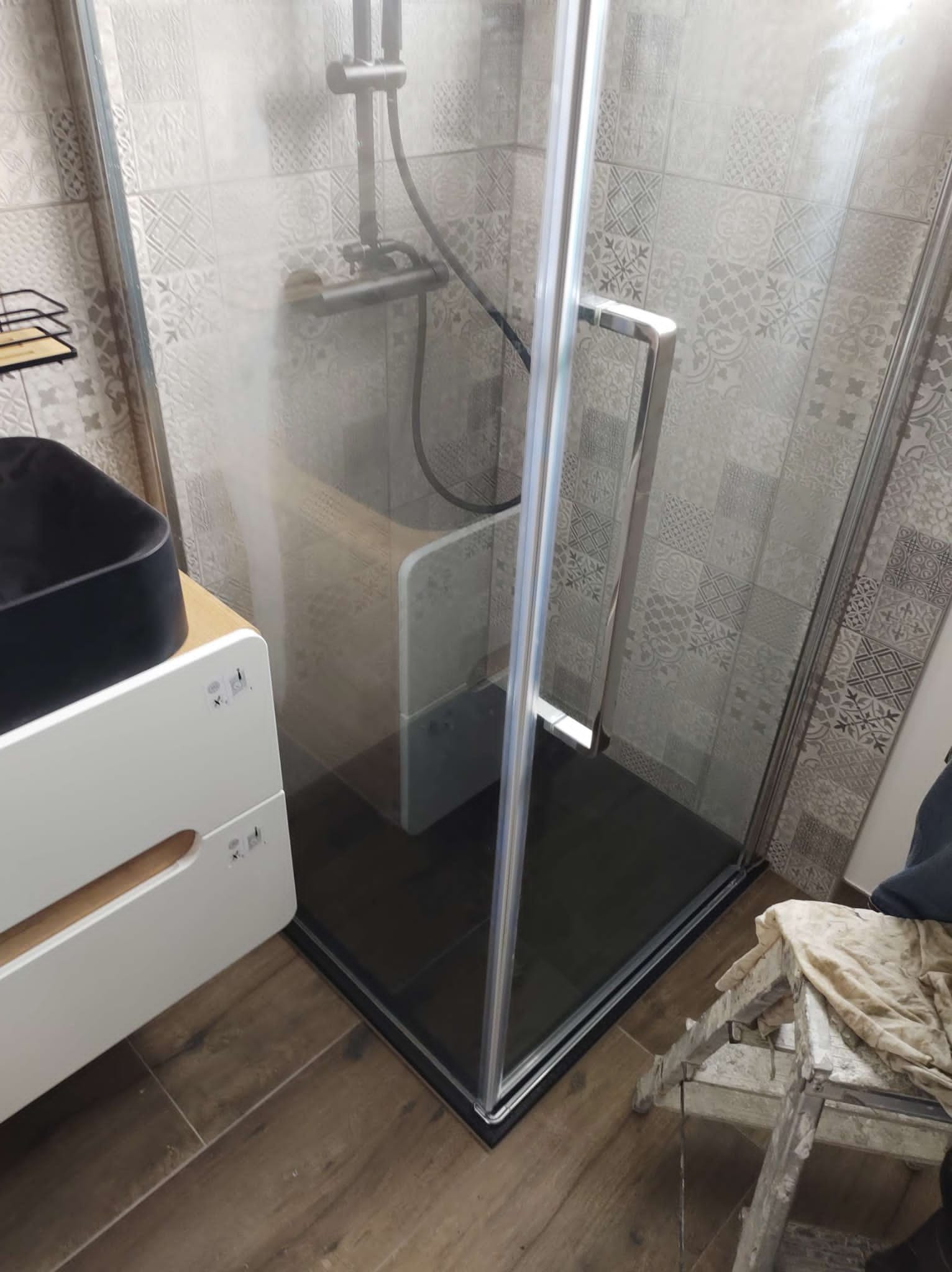 Nowoczesna kabina prysznicowa z chromowanymi profilami i czarnym brodzikiem, widoczna na tle płytek w patchworkowy wzór, obok umywalka nablatowa w kolorze czarnym i drewnopodobne płytki podłogowe...