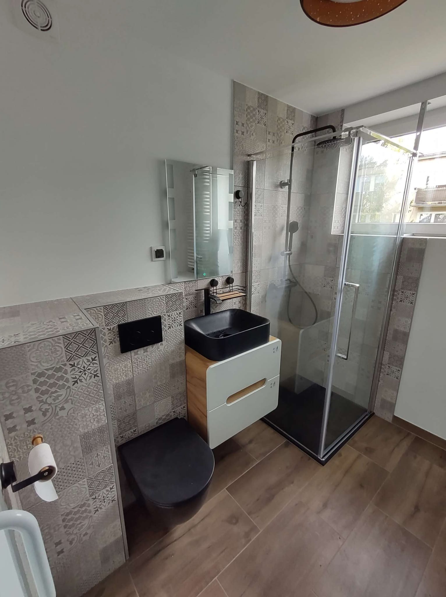 Nowoczesna łazienka z czarną umywalką nablatową, podwieszaną toaletą i kabiną prysznicową typu walk-in, wykończona płytkami w patchworkowy wzór oraz drewnopodobną podłogą.