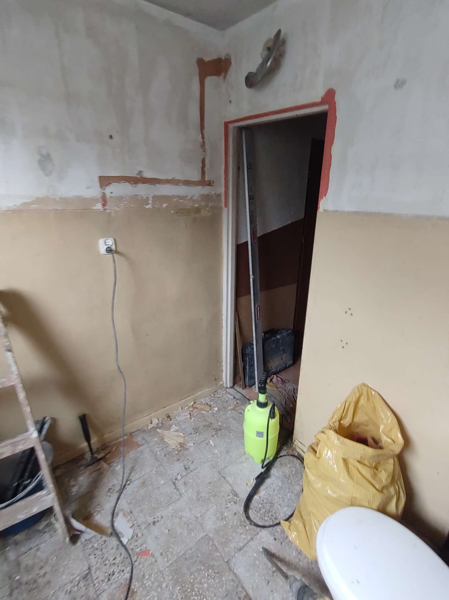 Pokój w trakcie przygotowania do remontu: ściany częściowo odrapane, widoczne ślady po instalacjach, drabina, narzędzia i worek z odpadami budowlanymi na podłodze.