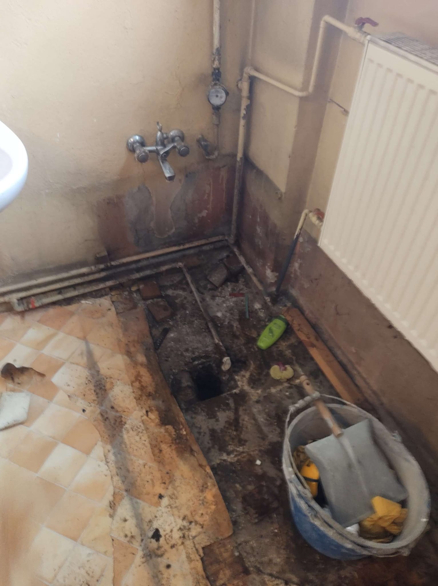 Demontaż starej podłogi w łazience, widoczne zerwane płytki, odsłonięta wylewka, wiadro z narzędziami i resztkami gruzu.