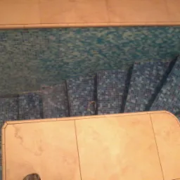 Mozaika w odcieniach błękitu i beżu pokrywająca schody i ścianę w pomieszczeniu. Widok z góry na fragment schodów z beżowym wykończeniem.